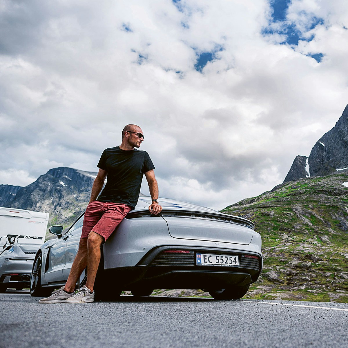 A man leans against a Porsche parked on the side of a road with snowy mountains in the background.