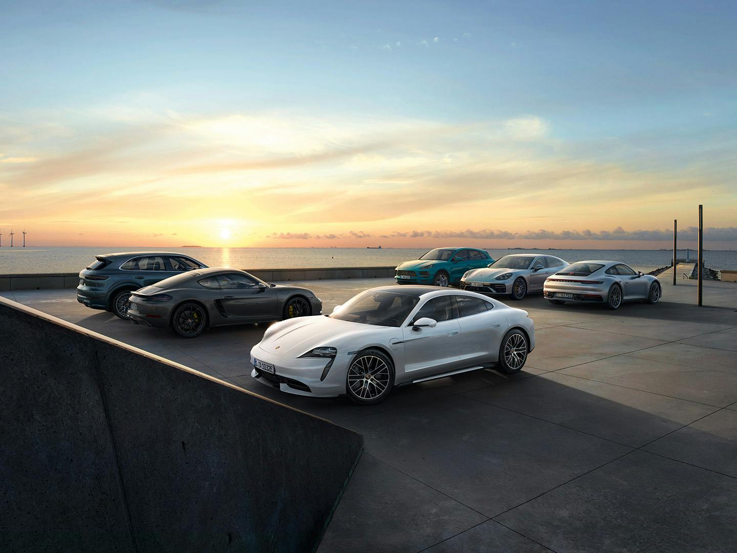 Eine Auswahl an Porsche-Modellen steht bei Sonnenuntergang an der Küste, darunter Taycan, 911, Panamera, Macan und Cayenne.