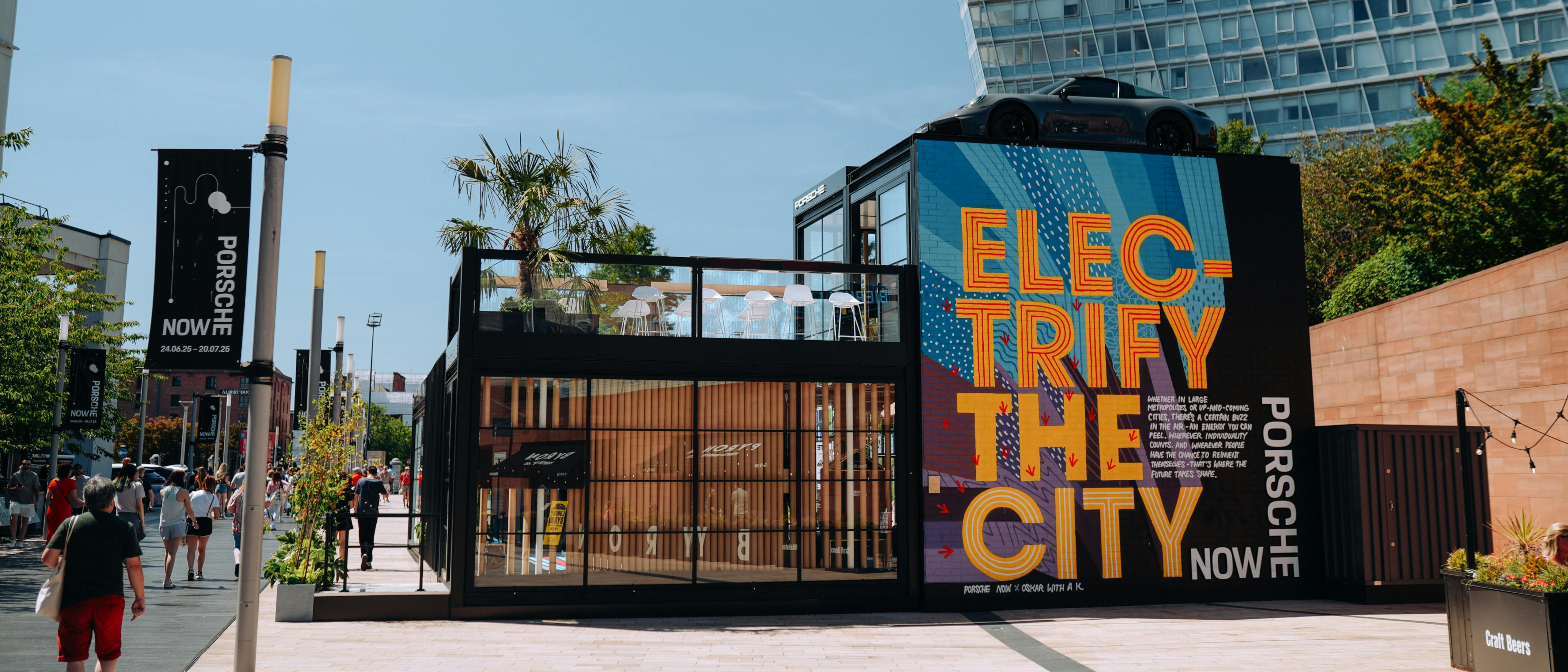 The Porsche Now pop-up in an urban plaza featuring a large sign reading “Electrify the City”, with a Porsche 911 displayed on the roof and people walking nearby.