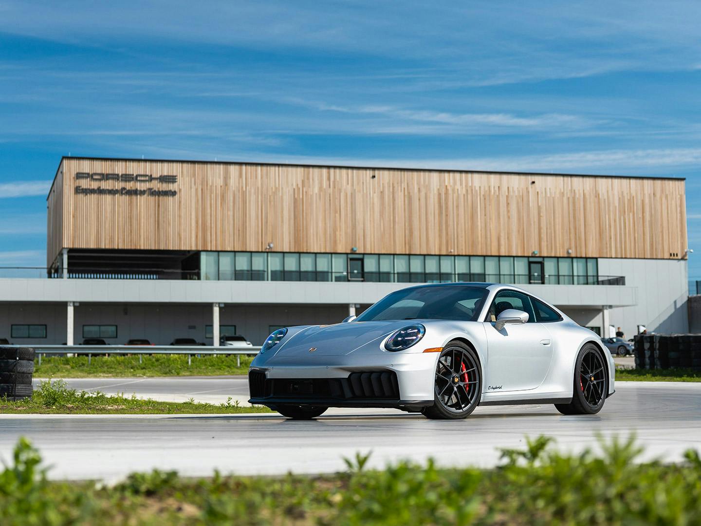 A silver Porsche 911 driving on a racetrack, with a modern building featuring a wood façade in the background.