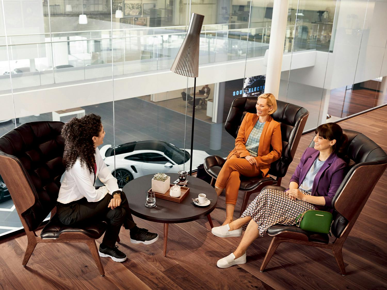 A female sales consultant talking with two clients in a relaxed setting inside a Porsche Centre. 