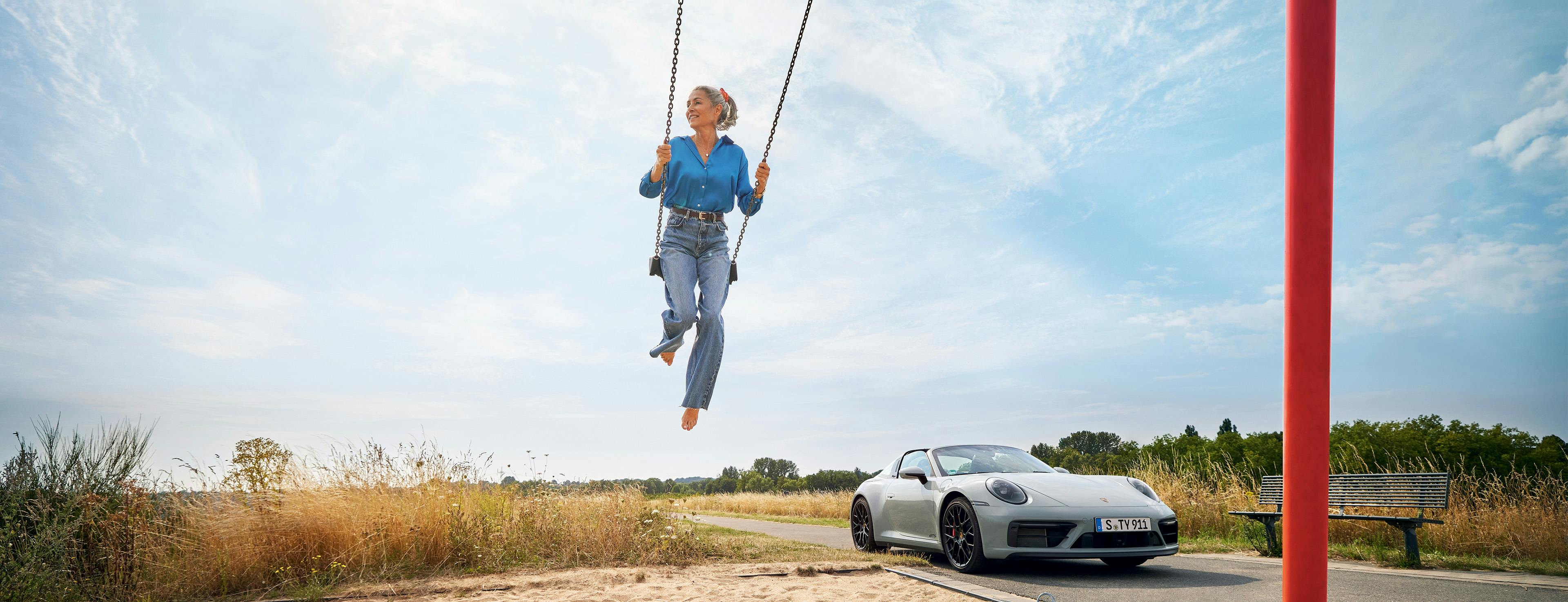 Woman playing on a swing in a sand park, while a Porsche 911 Targa parked by the side of the road.