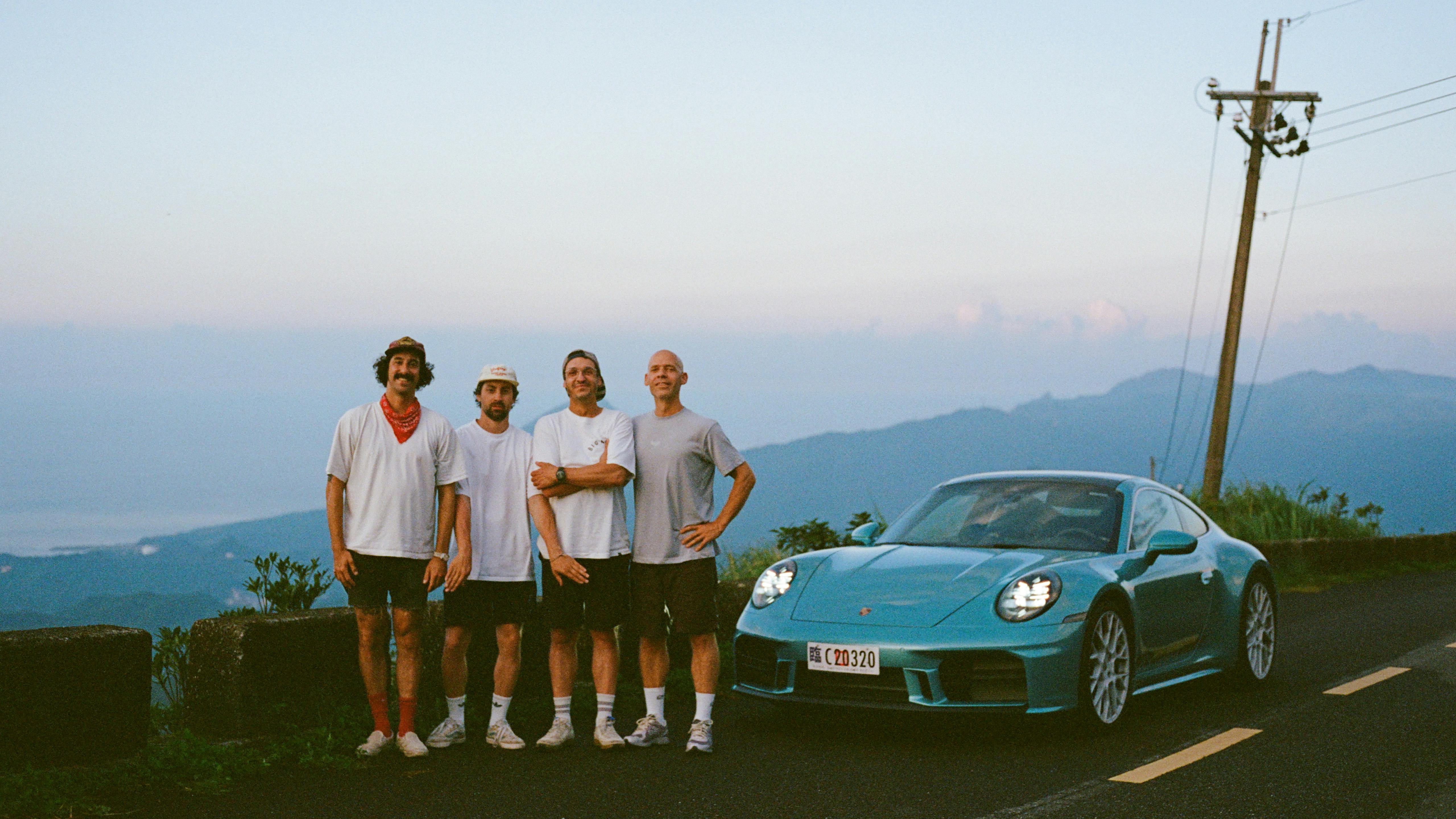 A Turquoise Porsche 911 parked on a mountain road beside four people, overlooking scenic hills and sky.