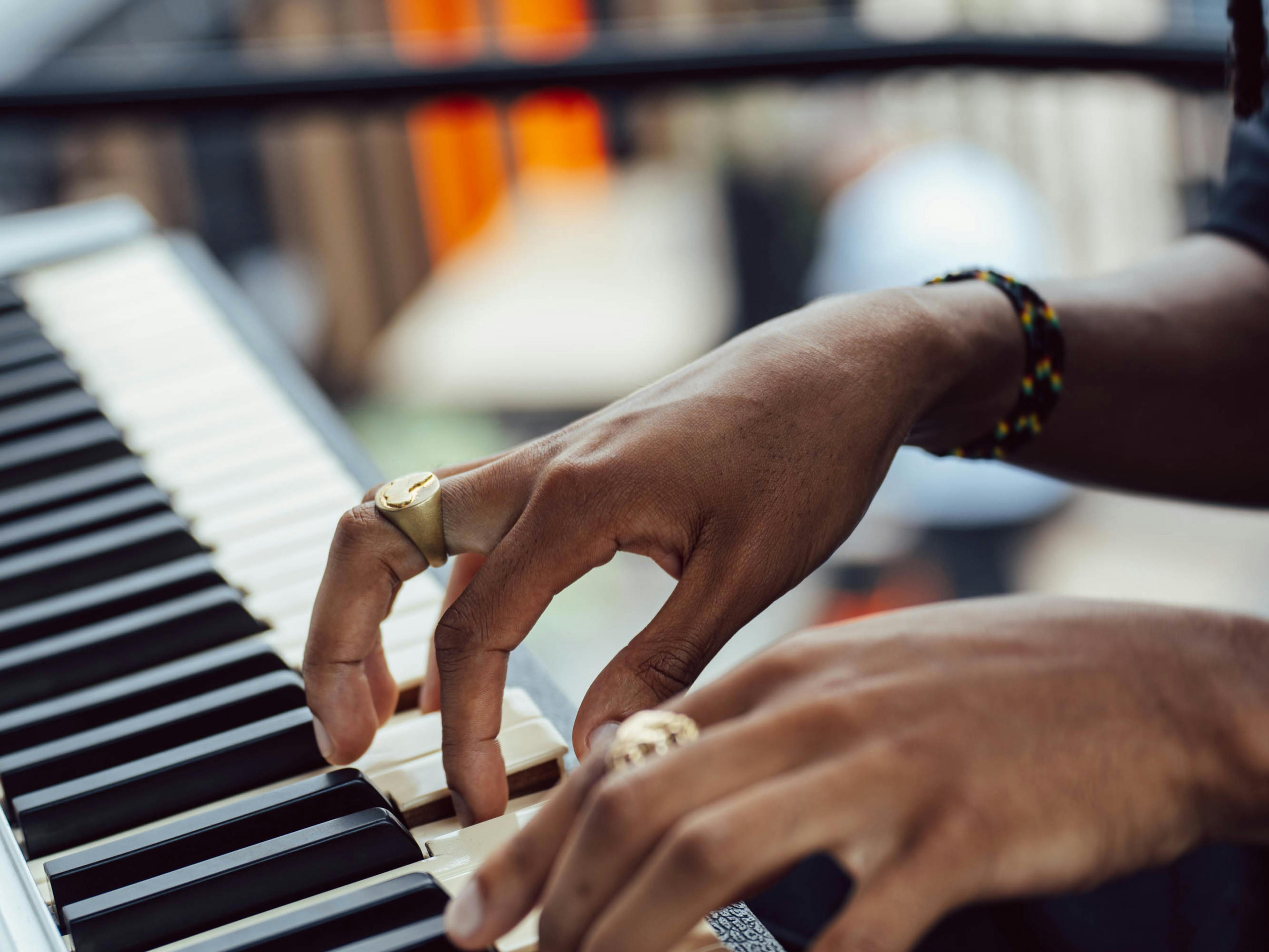 A close-up shot of a person’s hands with gold rings playing a keyboard. The background is blurred, showing hints of an outdoor event space. ​