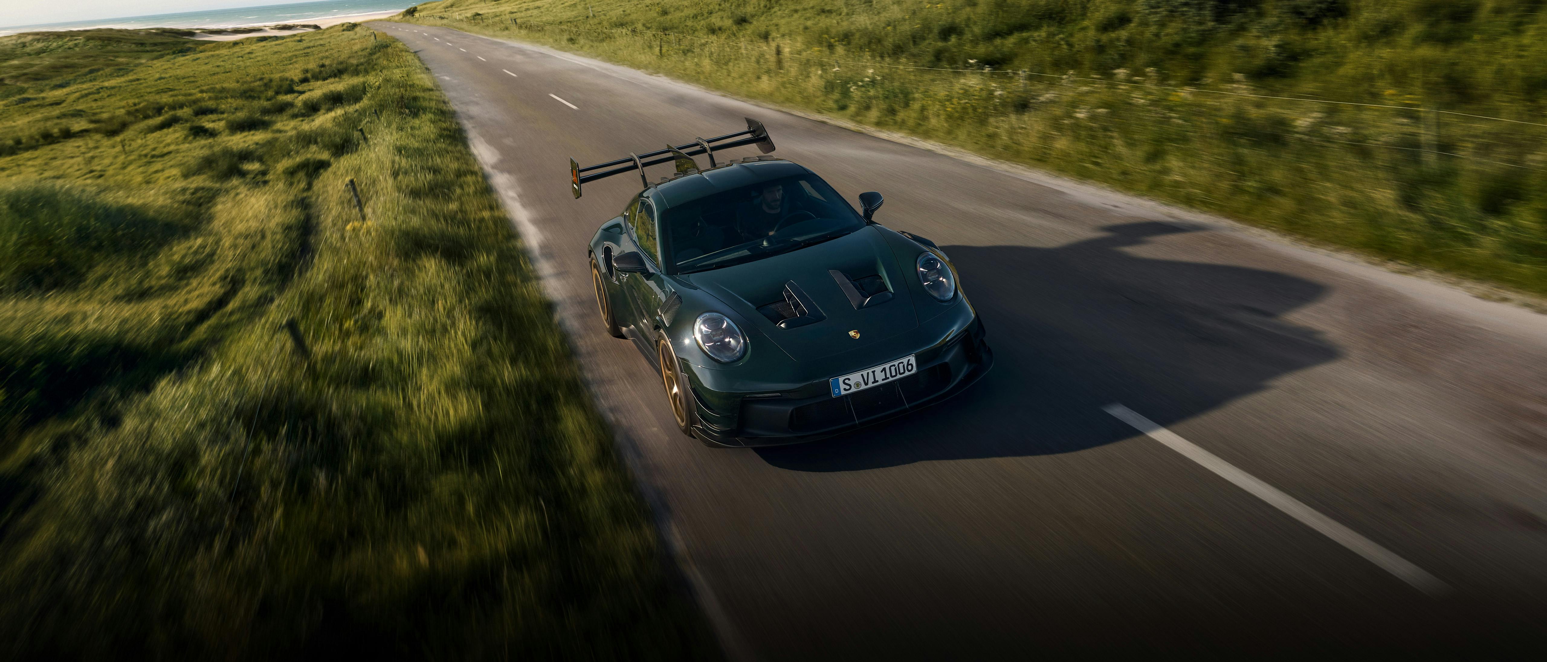 A GT3RS with Manthey kit in brewstergreen drives along a paved road between the dunes.