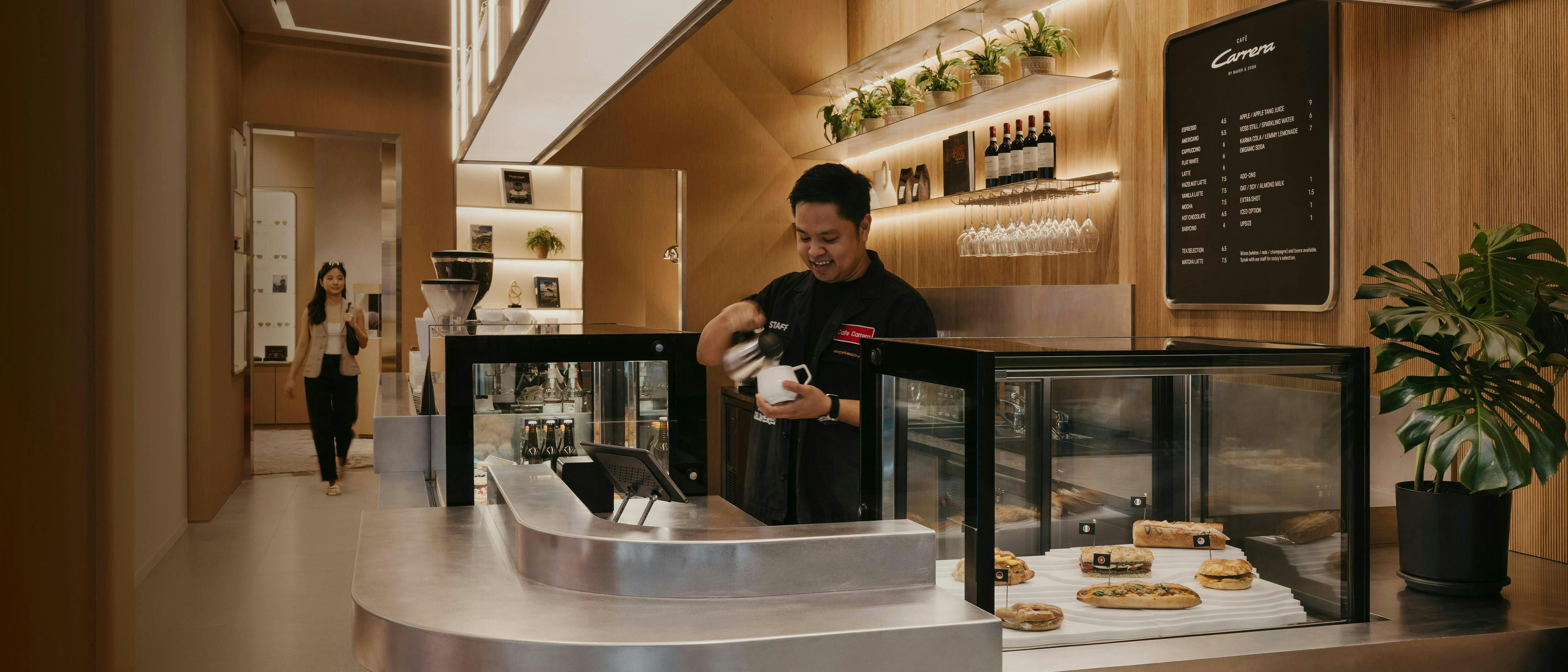 Barista at Café Carrera prepares coffee behind a modern counter  with sandwiches, wine display and warm wood interior.