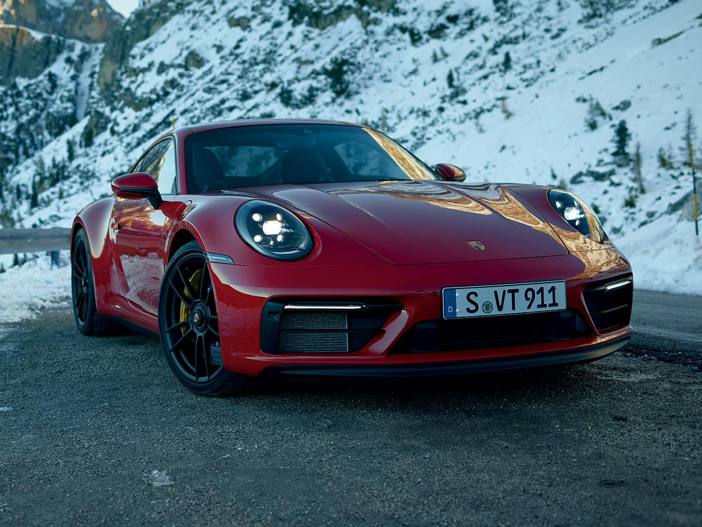 Une 911 Carrera GTS rouge sur une route de montagne en hiver, avec des sommets enneigés visibles en arrière-plan.