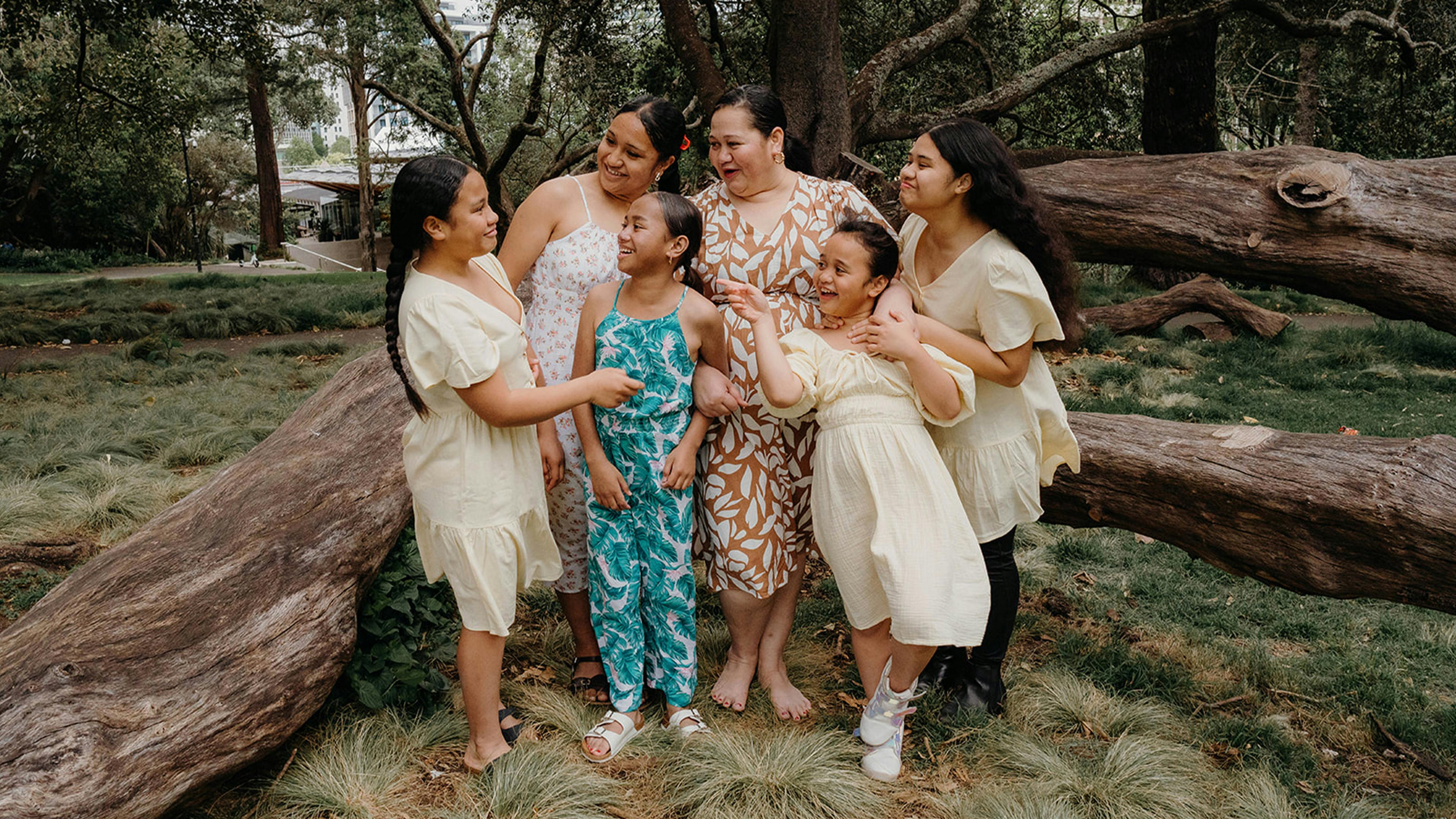 A group of six people, appearing to be a family, smiling and posing together outdoors in front of a large tree trunk. The individuals are wearing a mix of casual and patterned clothing.