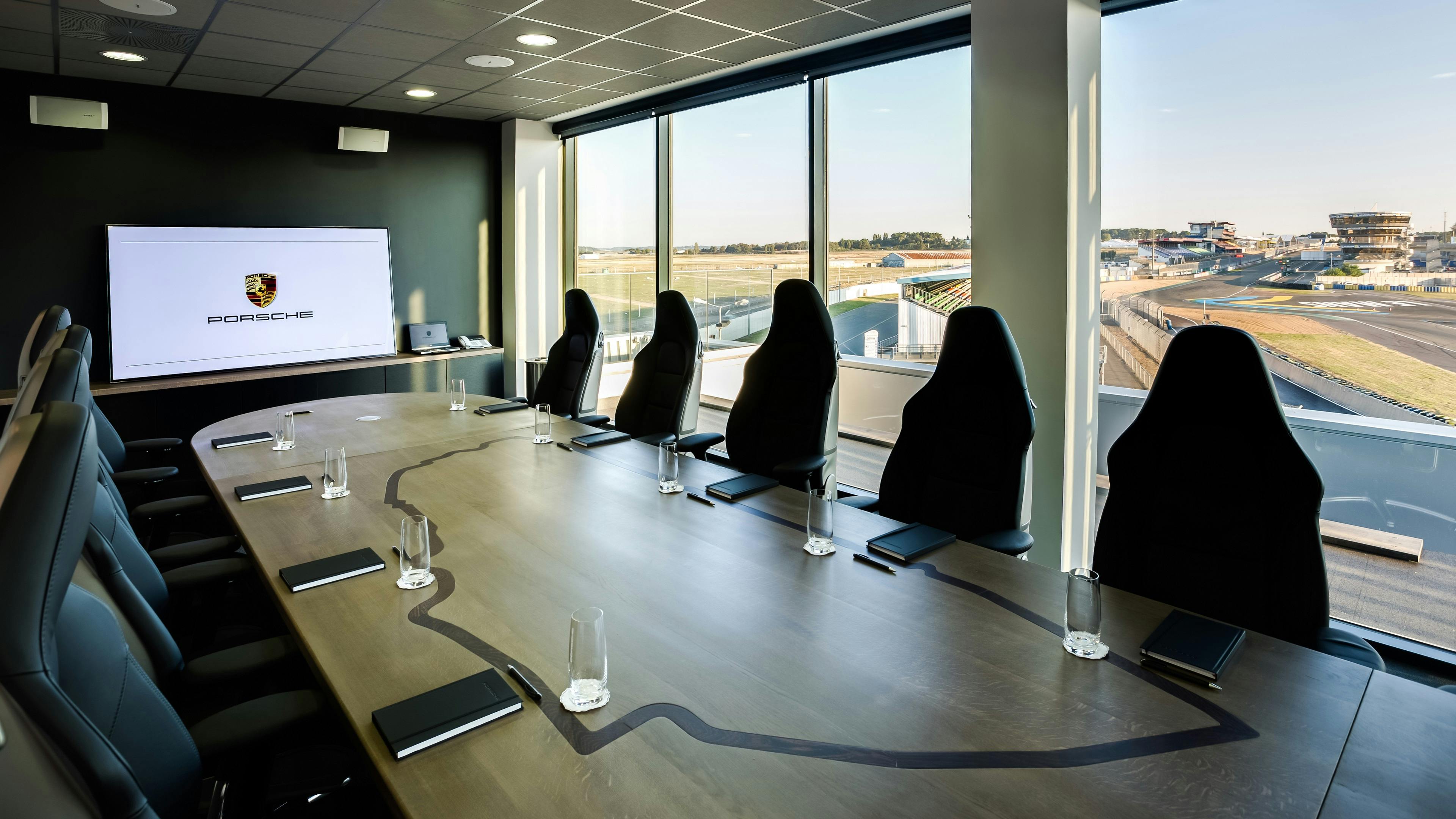 Salon Le Mans, avec la table sur mesure avec le circuit du Mans gravé dessus et autour 10 sièges de 911 faisant office de siège de bureau, vue sur le circuit Bugatti et la ligne droite des stands.
