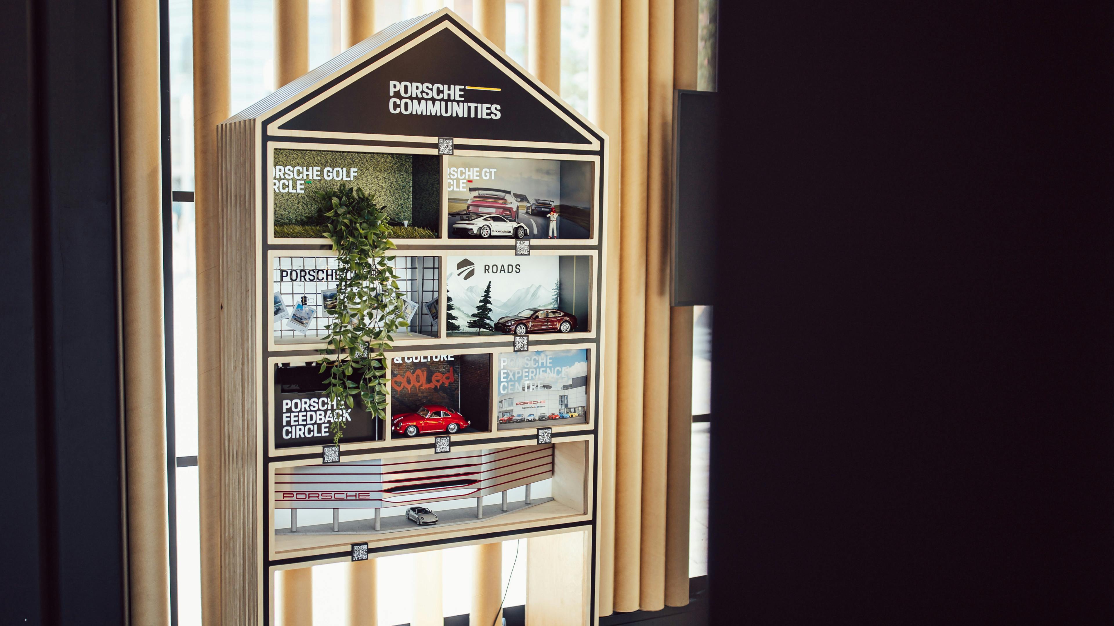 A house-shaped wooden display shelf titled “PORSCHE COMMUNITIES”. The compartments contain mini Porsche models.