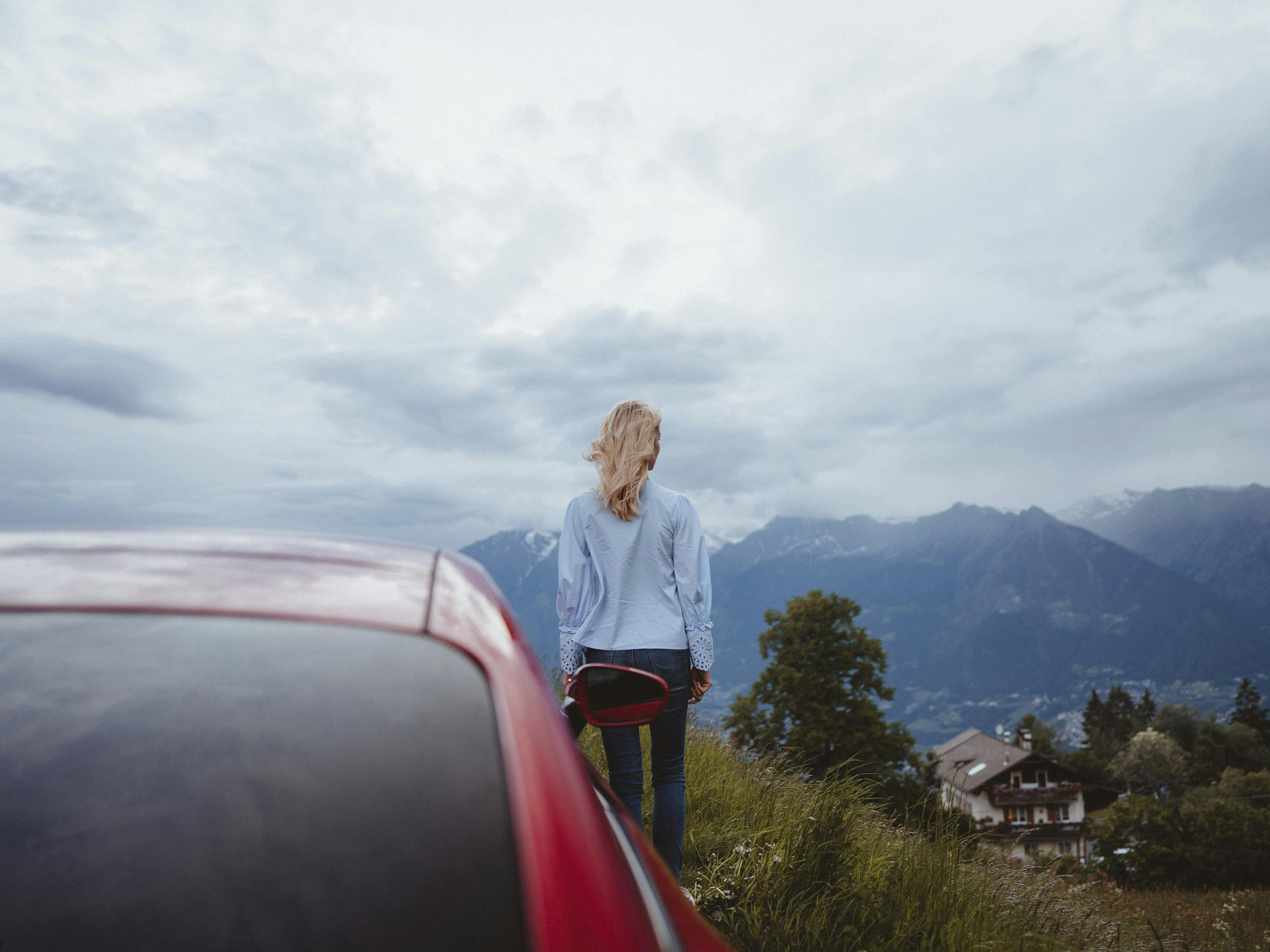 Frau mit blonden Haaren und hellblauer Bluse steht neben einem roten Auto auf einer Wiese und blickt in Richtung Berge. Im Hintergrund sind Häuser, Bäume und eine bewölkte Berglandschaft zu sehen.