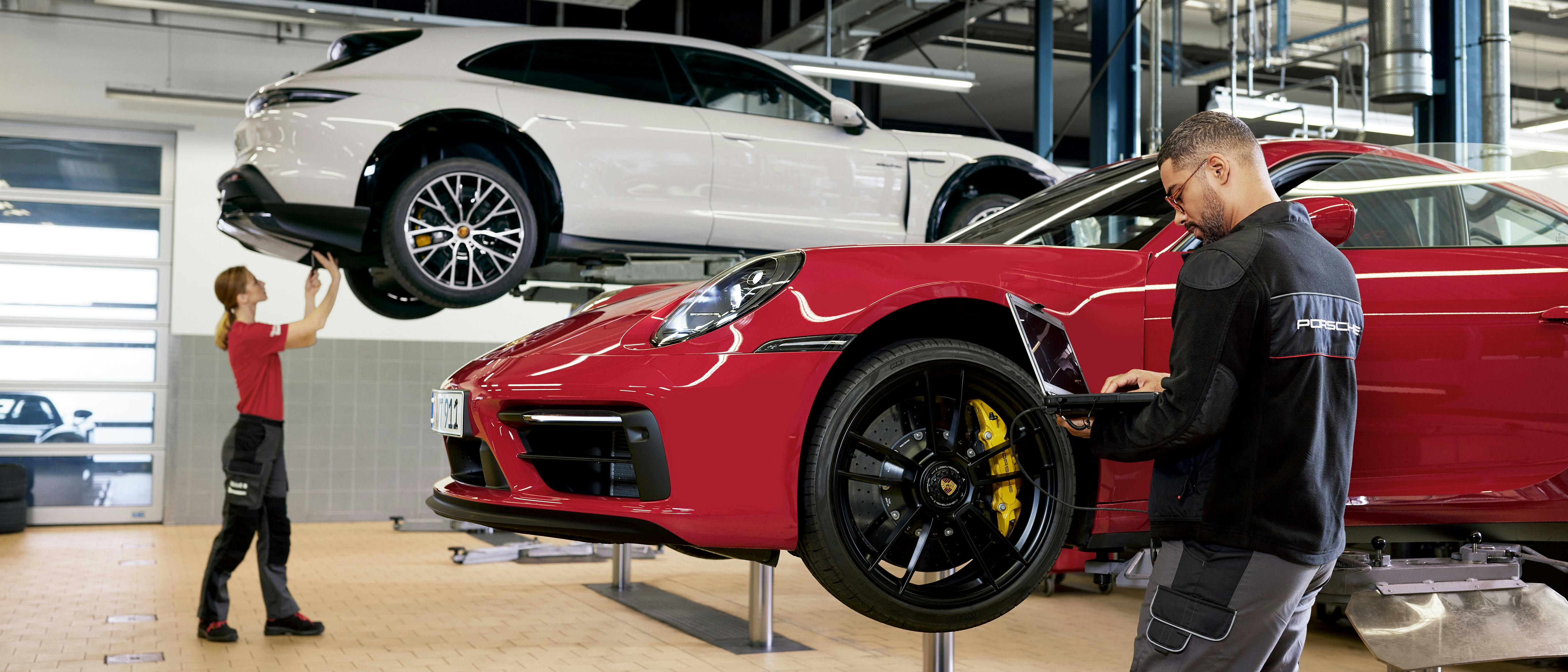 Een werkplaats met twee monteurs die aan twee Porsche auto's werken. Een man in een donker shirt met een Porsche logo erop, leunt over de voorkant van een rode auto en kijkt naar een laptop. Achter hem, aan de linkerkant, staat een vrouw in een rood shirt bij een witte Porsche Taycan die op een hefplatform staat.