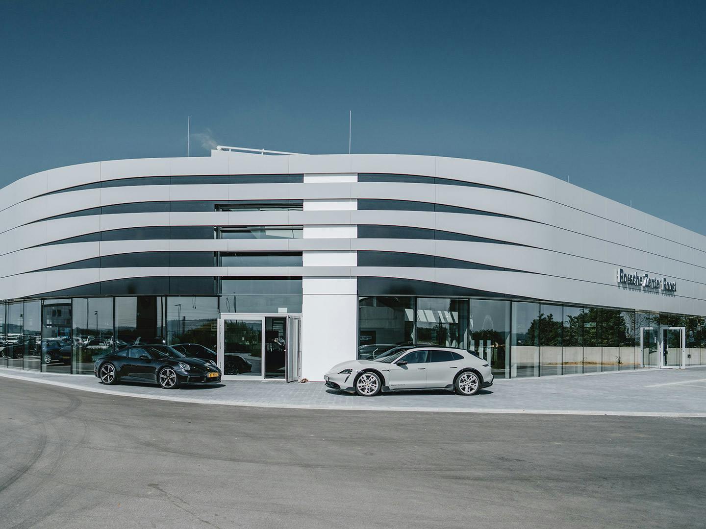 Extérieur du Porsche Zenter Lëtzebuerg de jour, avec de grandes lettres rouges Porsche sur un côté au-dessus de la façade vitrée continue, un panneau vertical blanc avec le logo Porsche noir et rouge à côté de l'entrée au centre, et plusieurs voitures Porsche stationnées devant le bâtiment.
