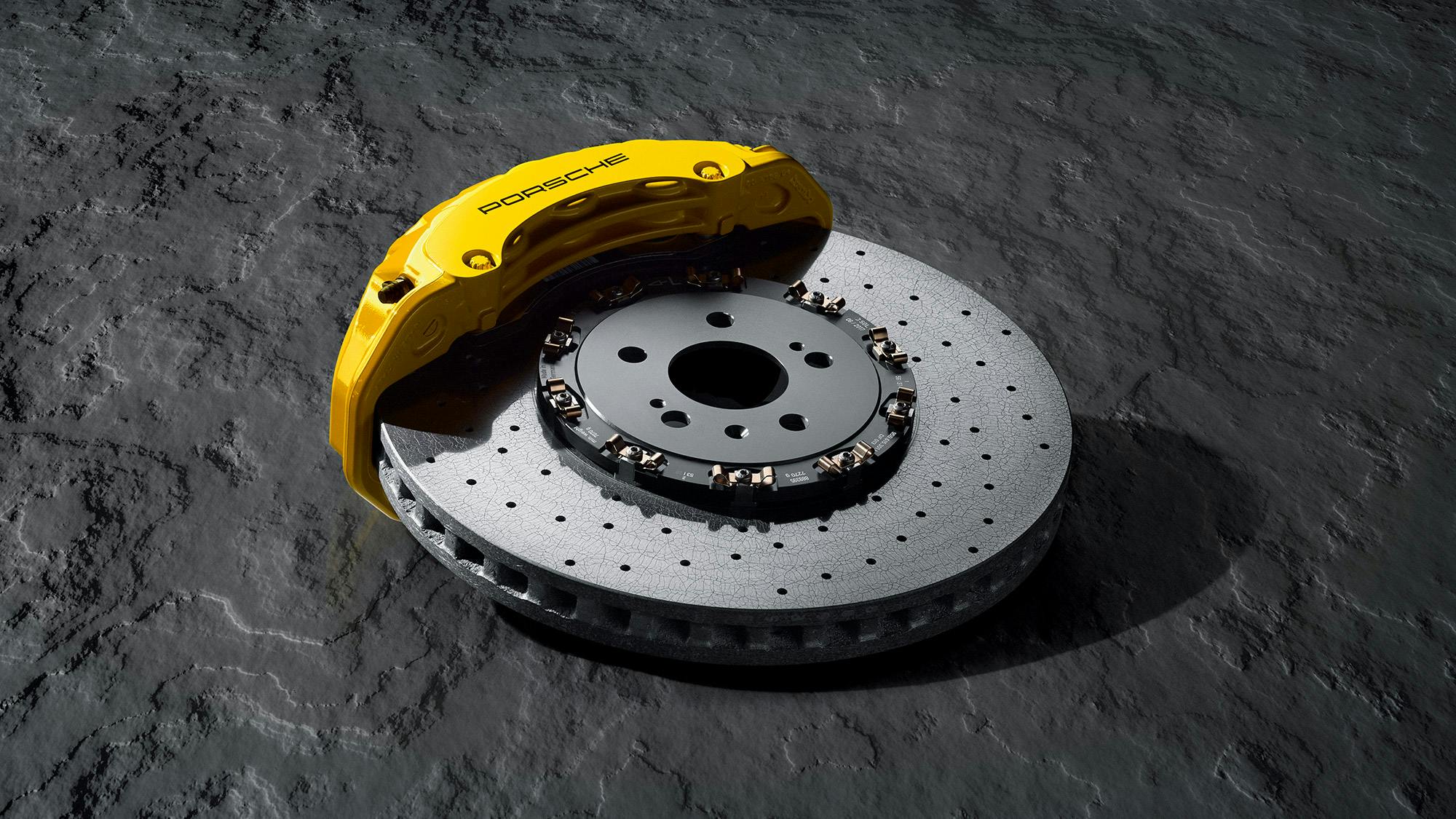 A yellow Porsche brake calliper and a perforated brake disc lie on a textured concrete slab.