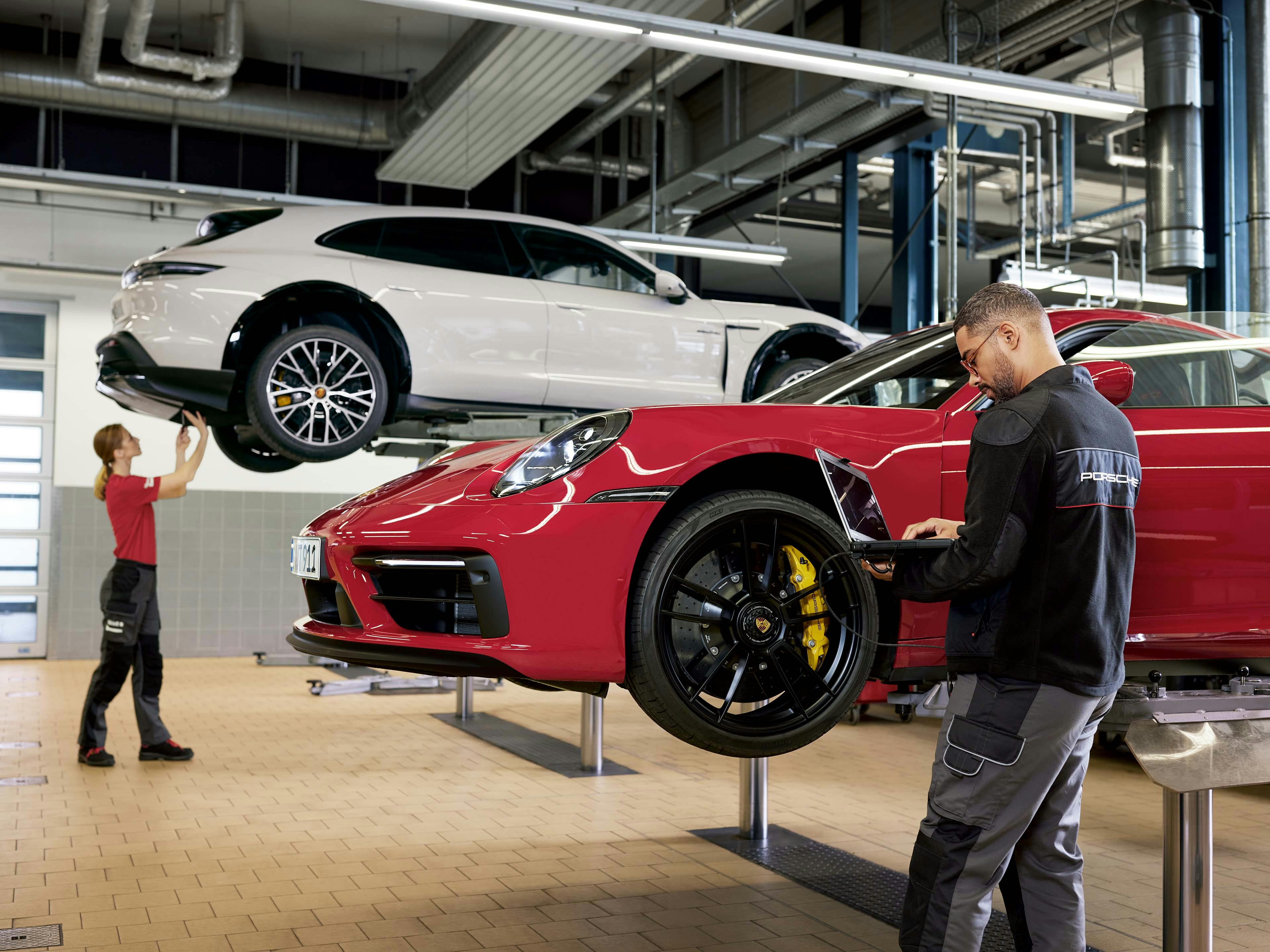 Een werkplaats met twee monteurs die aan twee Porsche auto's werken. Een man in een donker shirt met een Porsche logo erop, leunt over de voorkant van een rode auto en kijkt naar een laptop. Achter hem, aan de linkerkant, staat een vrouw in een rood shirt bij een witte Porsche Taycan die op een hefplatform staat.