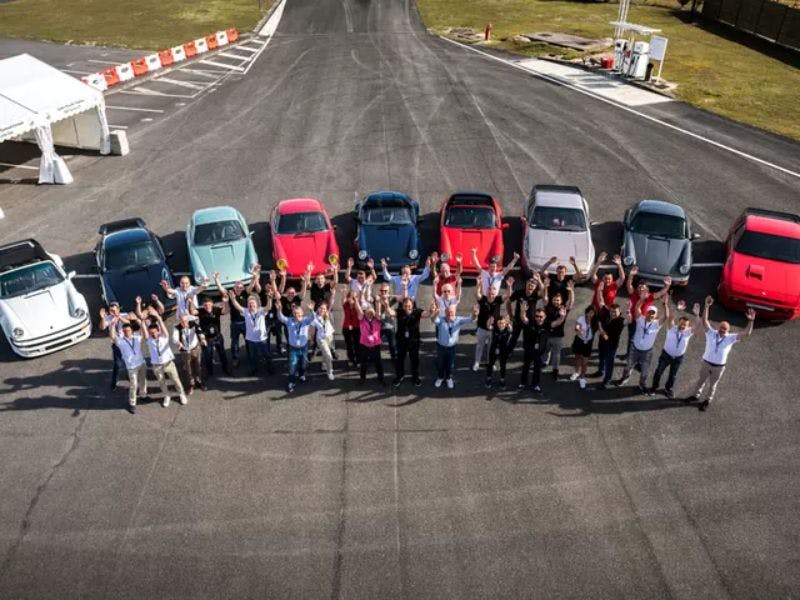 Les participants au Concours de Restauration se tiennent sur le circuit. Derrière eux, les neuf voitures participantes sont alignées les unes à côté des autres.