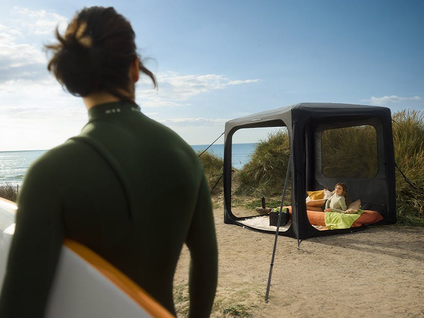 A Porsche pavilion set up in the dunes is viewed by a woman with a surfboard in her arms.