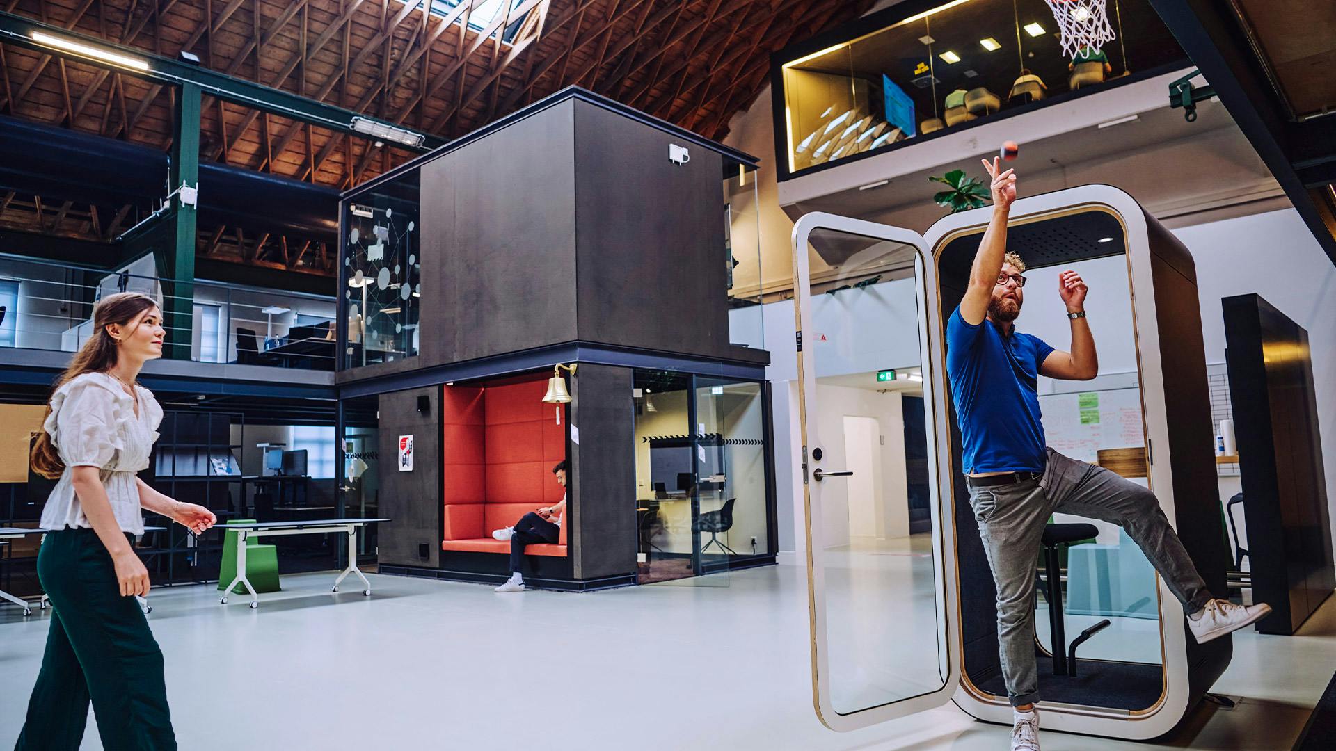 Moderne Arbeitsumgebung mit offener Architektur: Zwei Personen spielen Basketball an einem integrierten Korb in einem flexiblen Büro‑ und Kreativraum mit modularen Arbeitsbereichen, Sitzgelegenheiten und hoher Hallendecke.