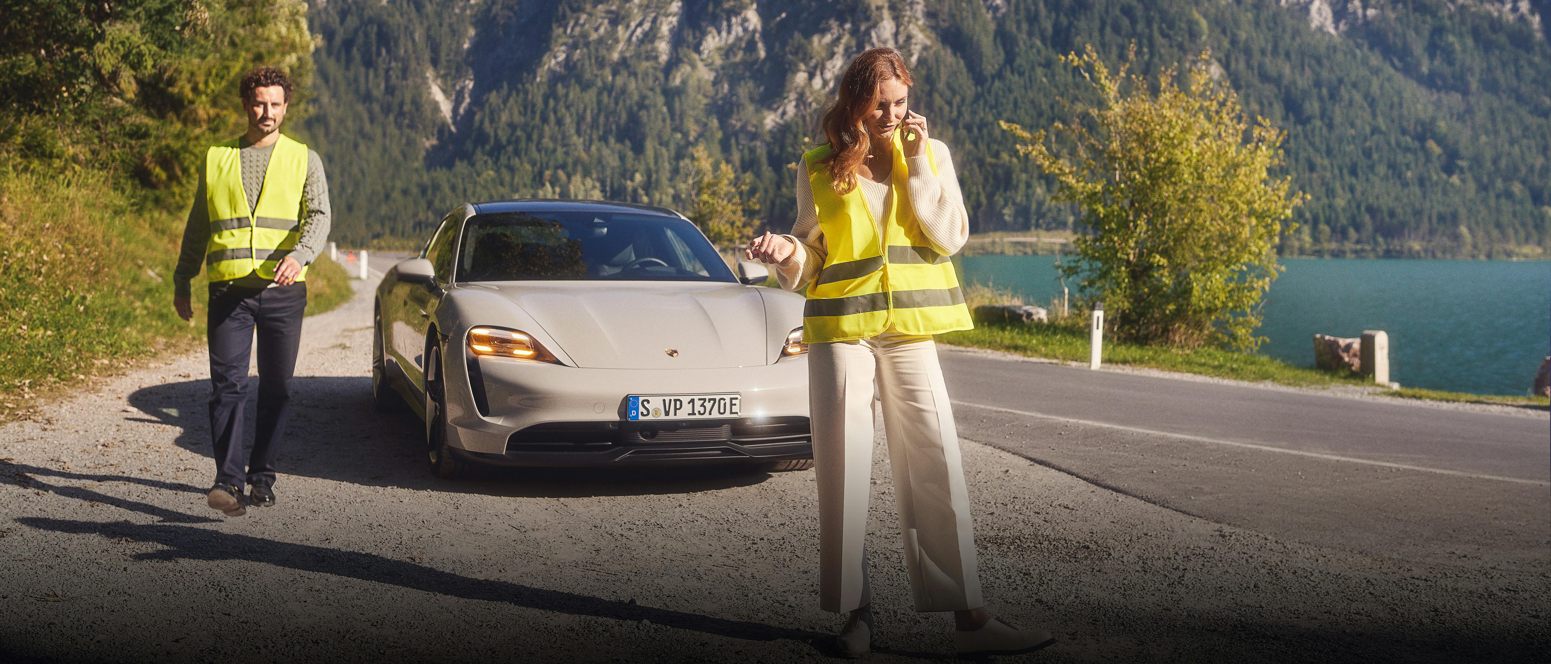 Due persone con giubbotti catarifrangenti: la donna parla al cellulare davanti a una Porsche Taycan, mentre un uomo cammina nella sua direzione.