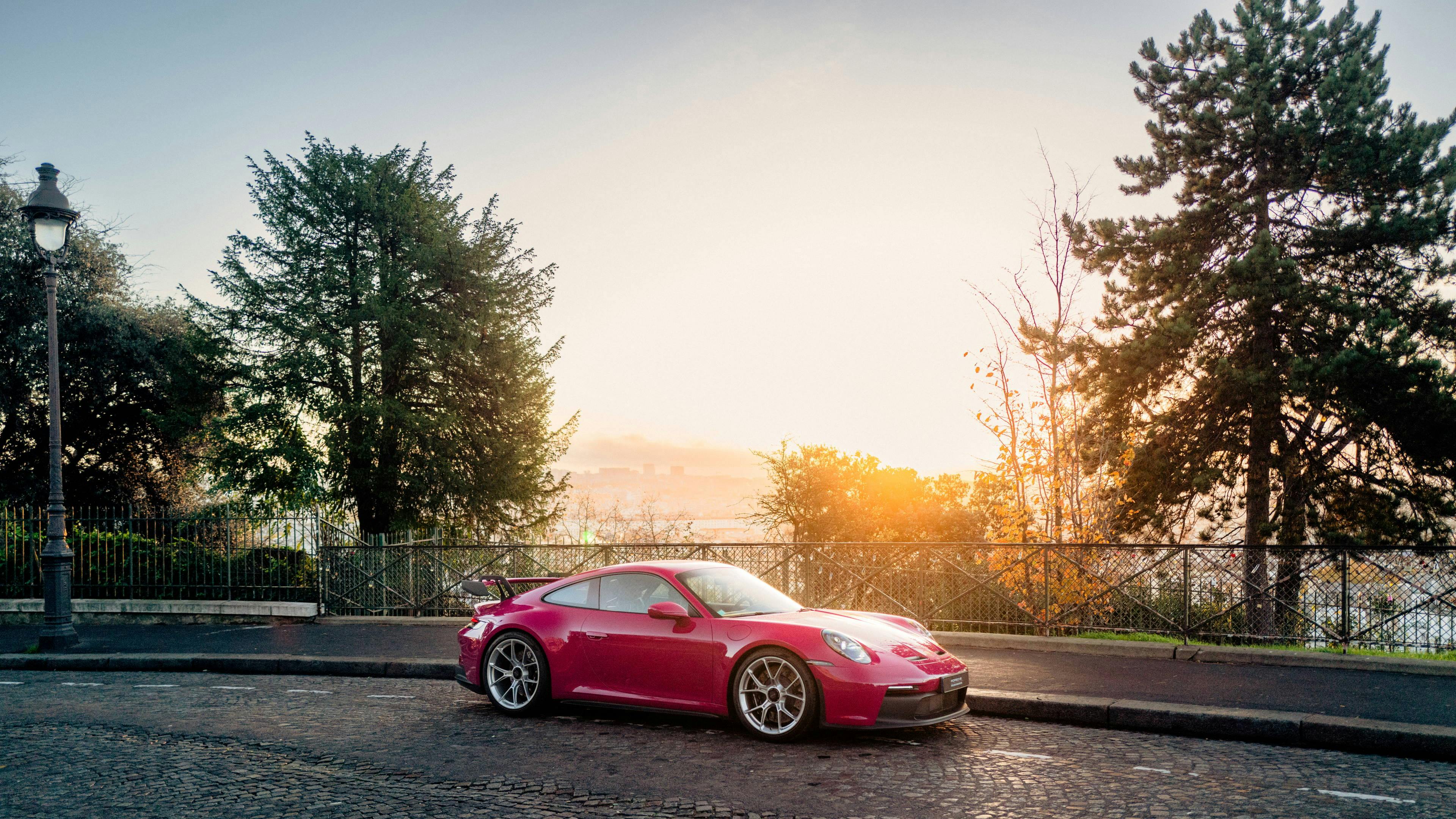 A red Porsche car is parked on a cobbled street, the sun is rising in the background.