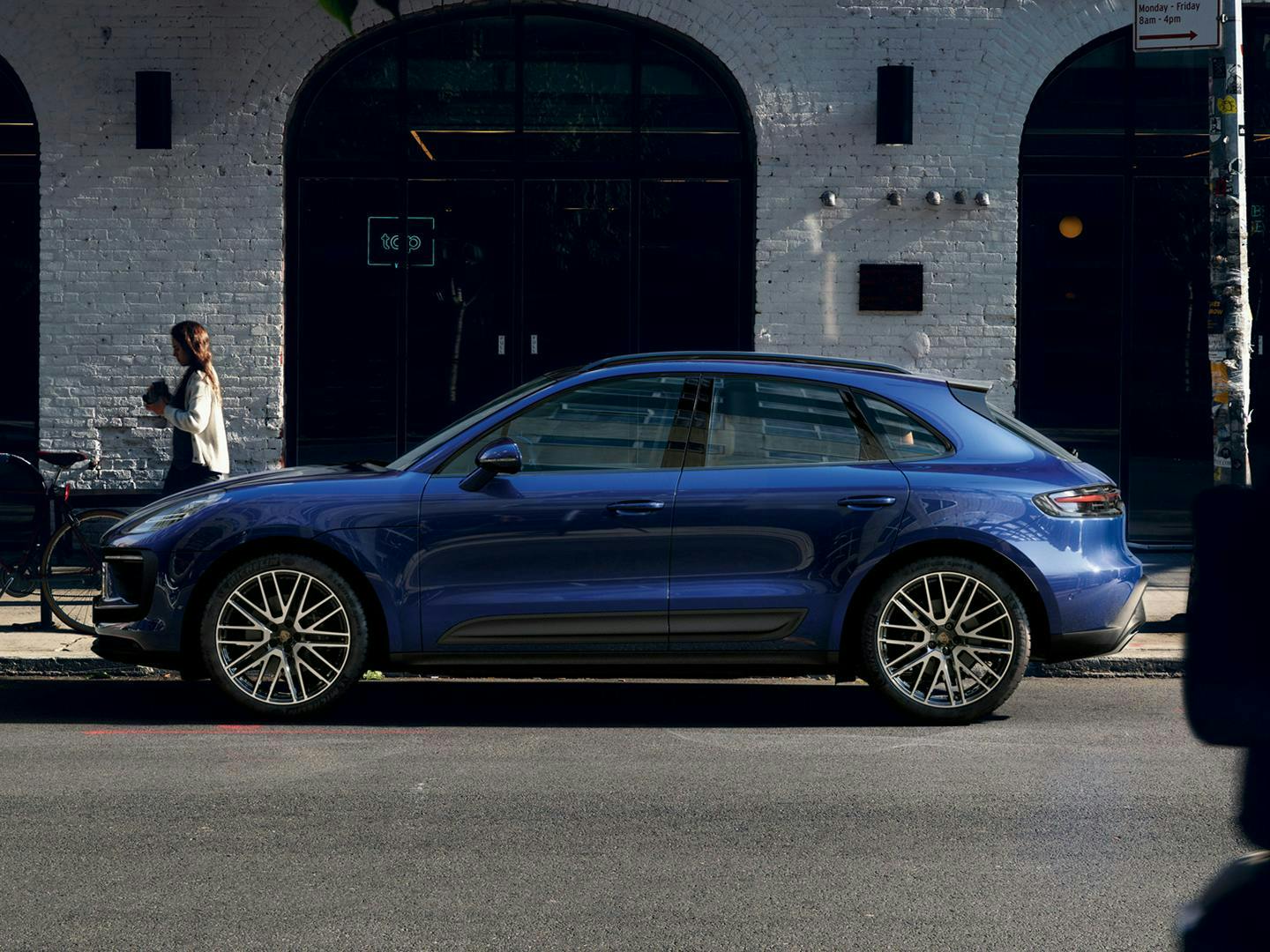 The Macan parked on the road, in front of a white building that looks like a shop, there is a man walking past in the foreground while a woman walking in the opposite direction in the foreground, nearer to the car.