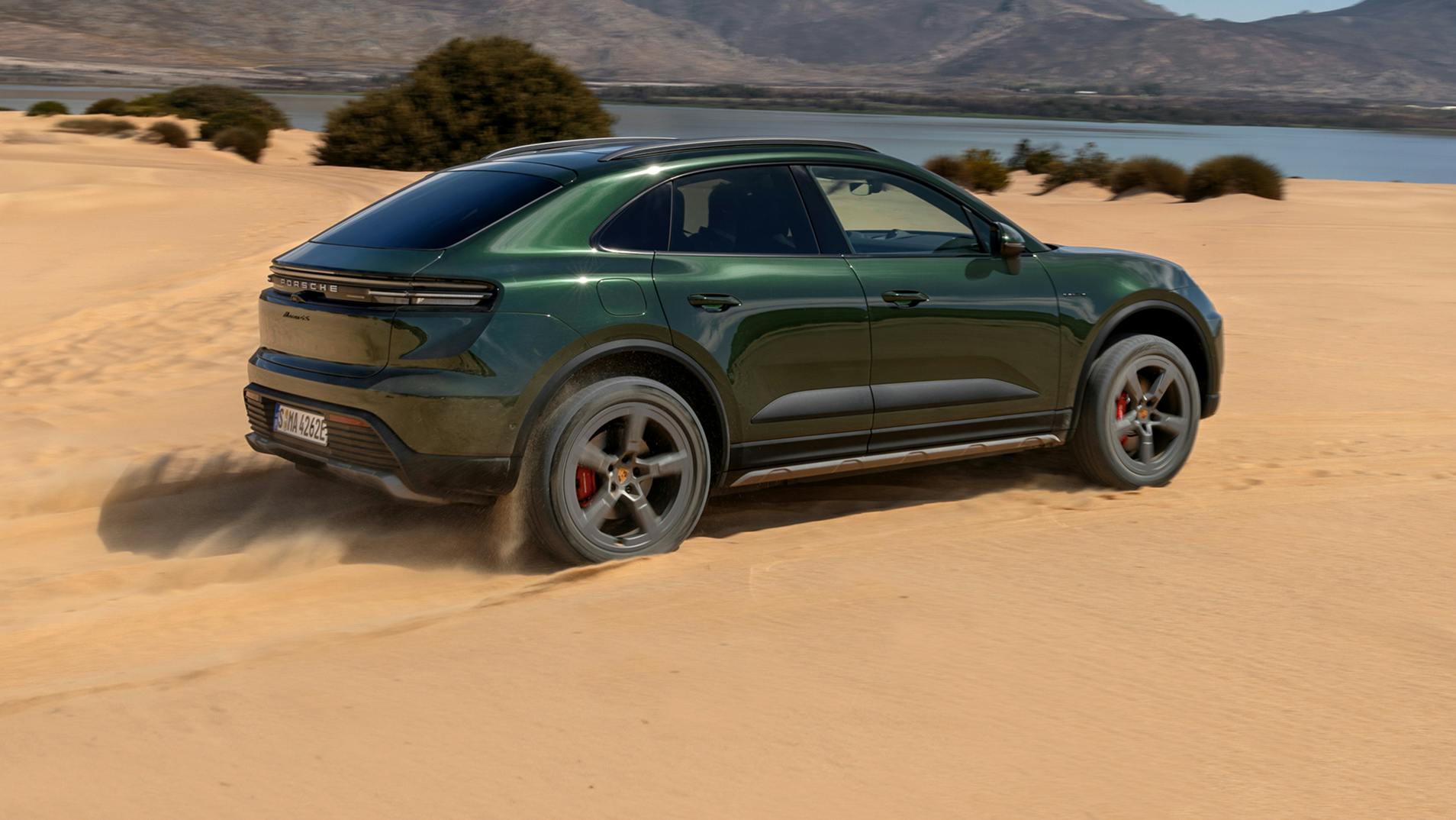 A Porsche Macan 4S driving over sand. A lake and a mountain range are in the background.