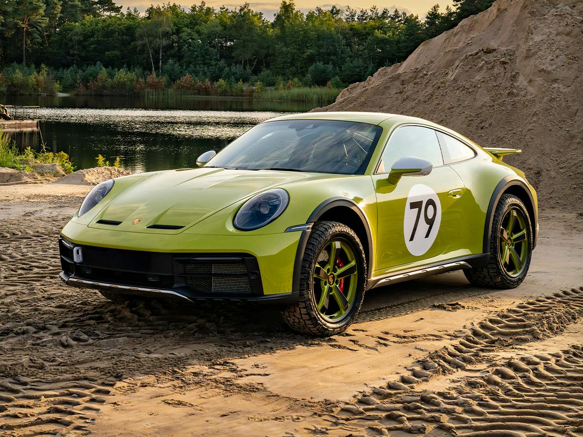 Porsche 911 Dakar in Paint to Sample colour Lindengreen with white lollipop number 79 on the door, featuring wheels with Lindgreen accents, parked on sandy terrain at sunset with trees and water in the background.