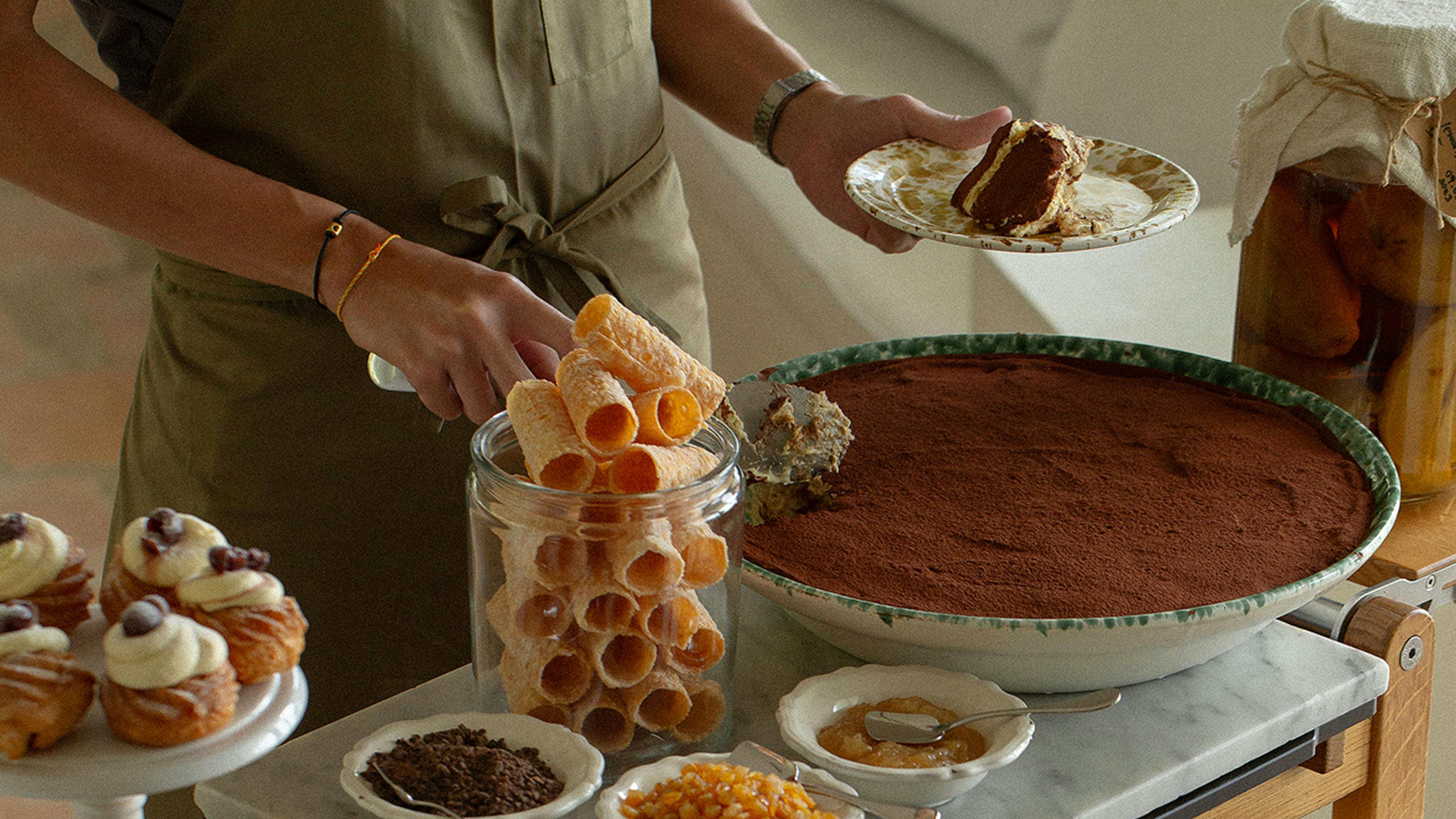 Wooden dessert cart with large tiramisu dish, jars of rolled wafers, toppings, and trays of pastries at Fico restaurant East Coast.