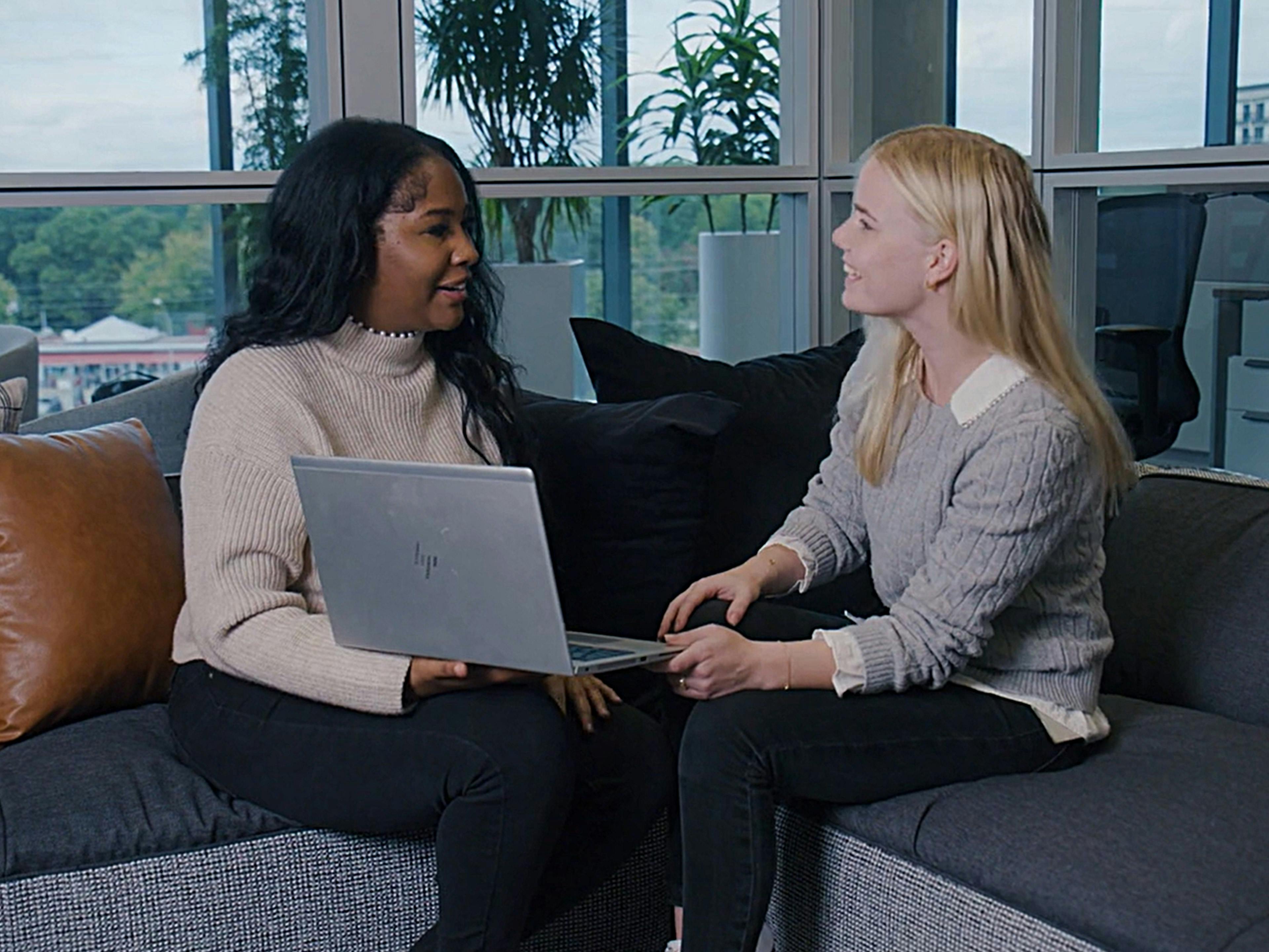Two women sit on a couch in an office setting with a laptop computer.