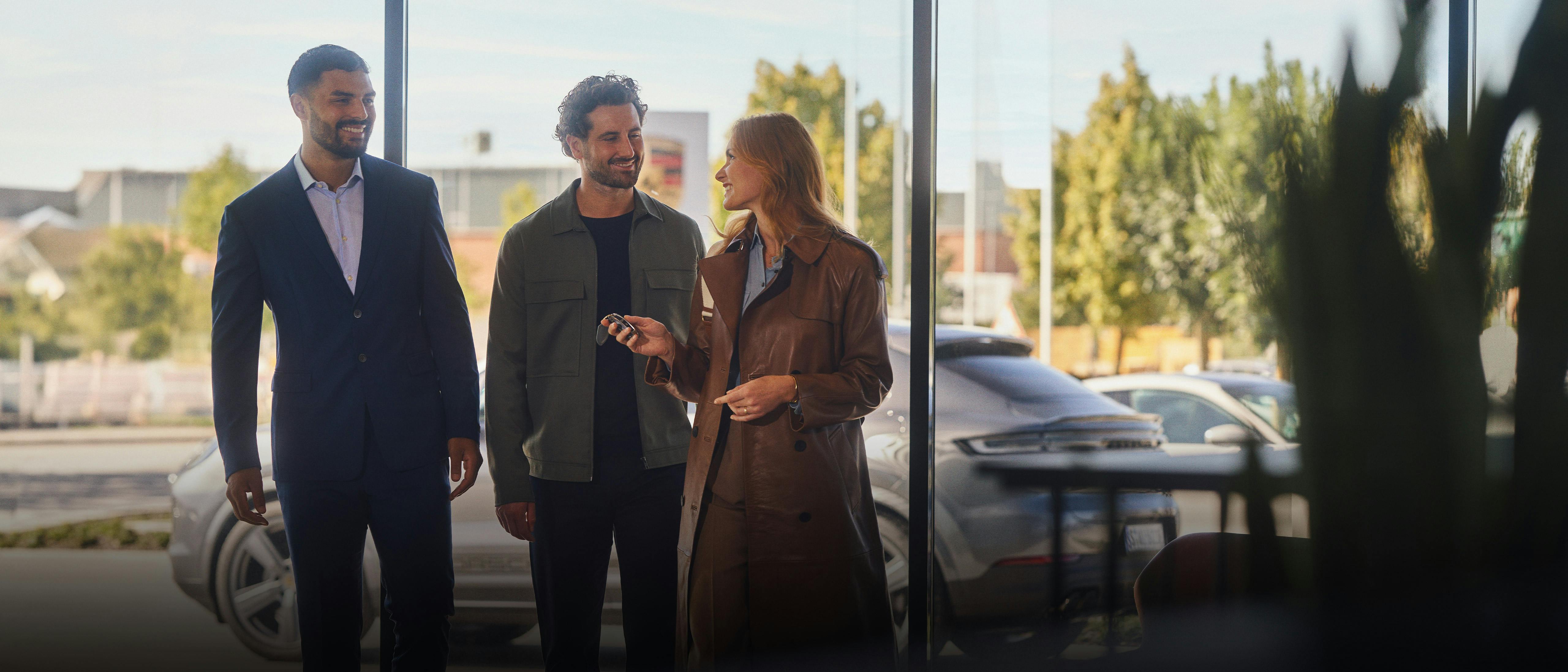 A couple is standing in a Porsche Center together with a consultant. The woman has a car key in her hand.