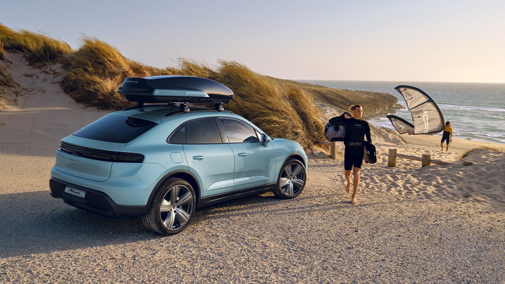A blue Porsche SUV parked on a sandy beach, equipped with a roof box. A man in a wetsuit walks nearby, holding gear, with kite surfers in the background.