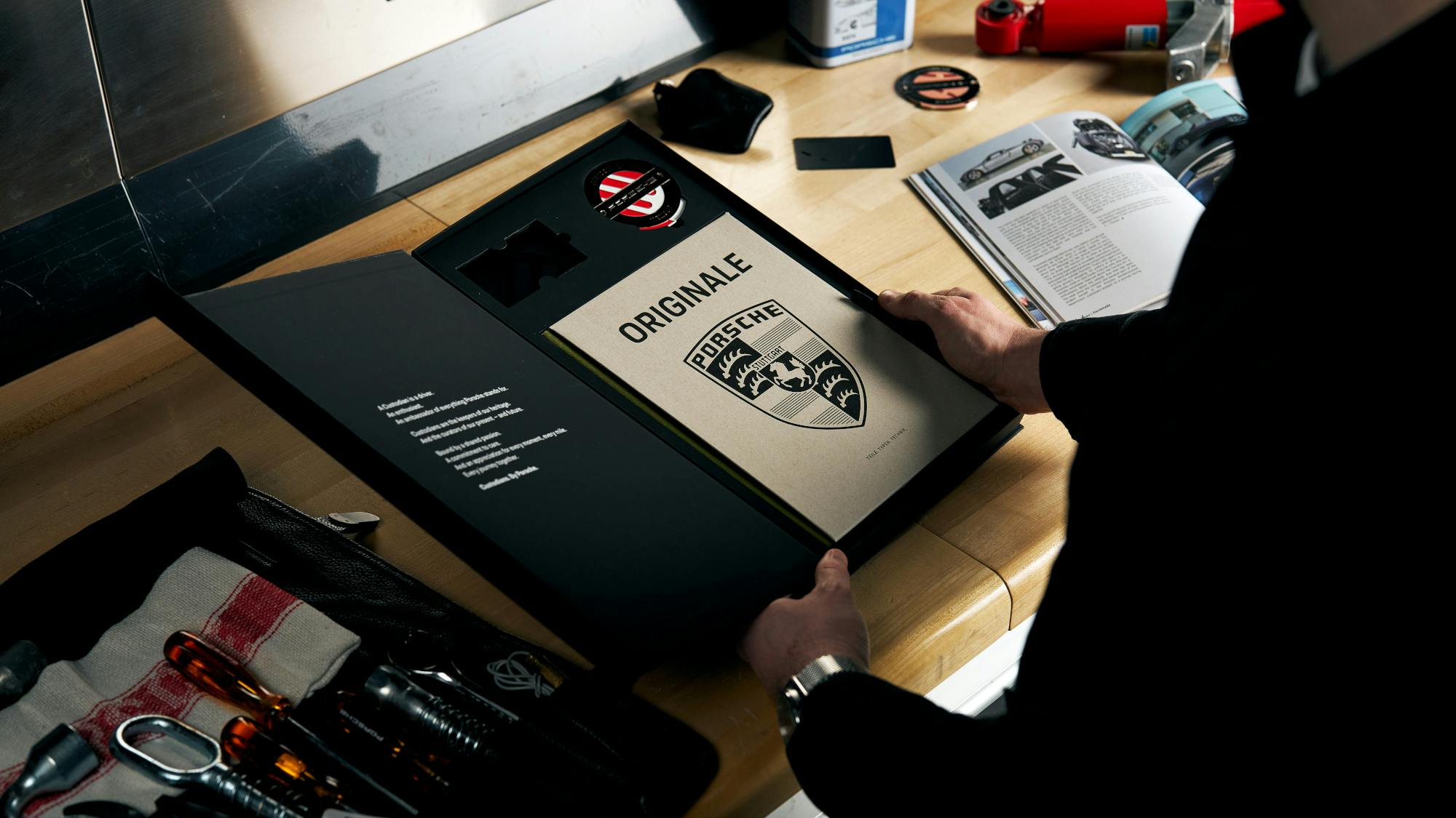 An overhead, side-angle view of a person wearing a black jacket and a silver watch, opening a matte black presentation box on a wooden workbench. The inside of the box cover features white text, while the main compartment holds a tan "ORIGINALE" Porsche booklet with a large black crest and a small round metallic badge. The cluttered workbench around the box includes a black tool roll with vintage tools, an open car magazine, and various mechanical parts under bright overhead lighting.