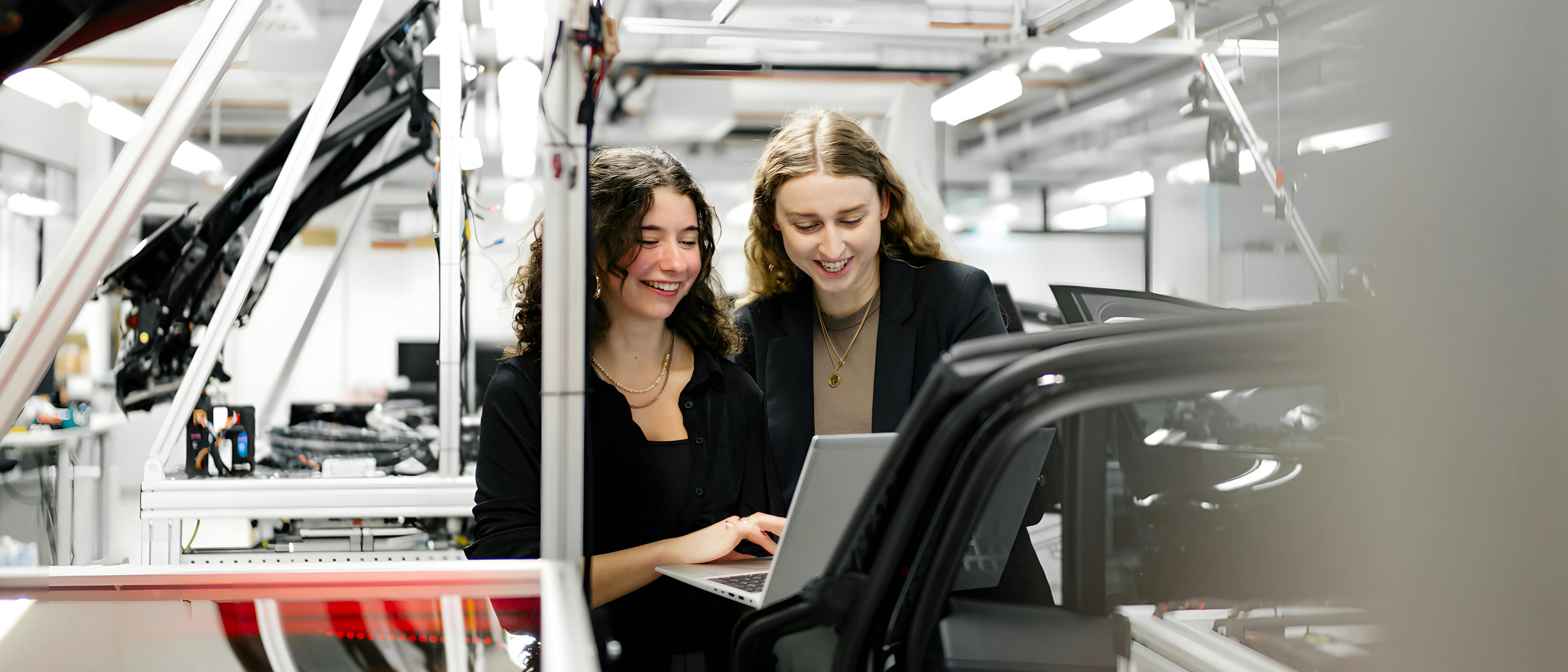 Zwei Studentinnen stehen in einer hellen, technischen Arbeitsumgebung neben einem Fahrzeug und betrachten gemeinsam einen Laptop. Im Vordergrund sind Fahrzeugrahmen und Montageelemente zu sehen, im Hintergrund industrielle Anlagen und Beleuchtung.