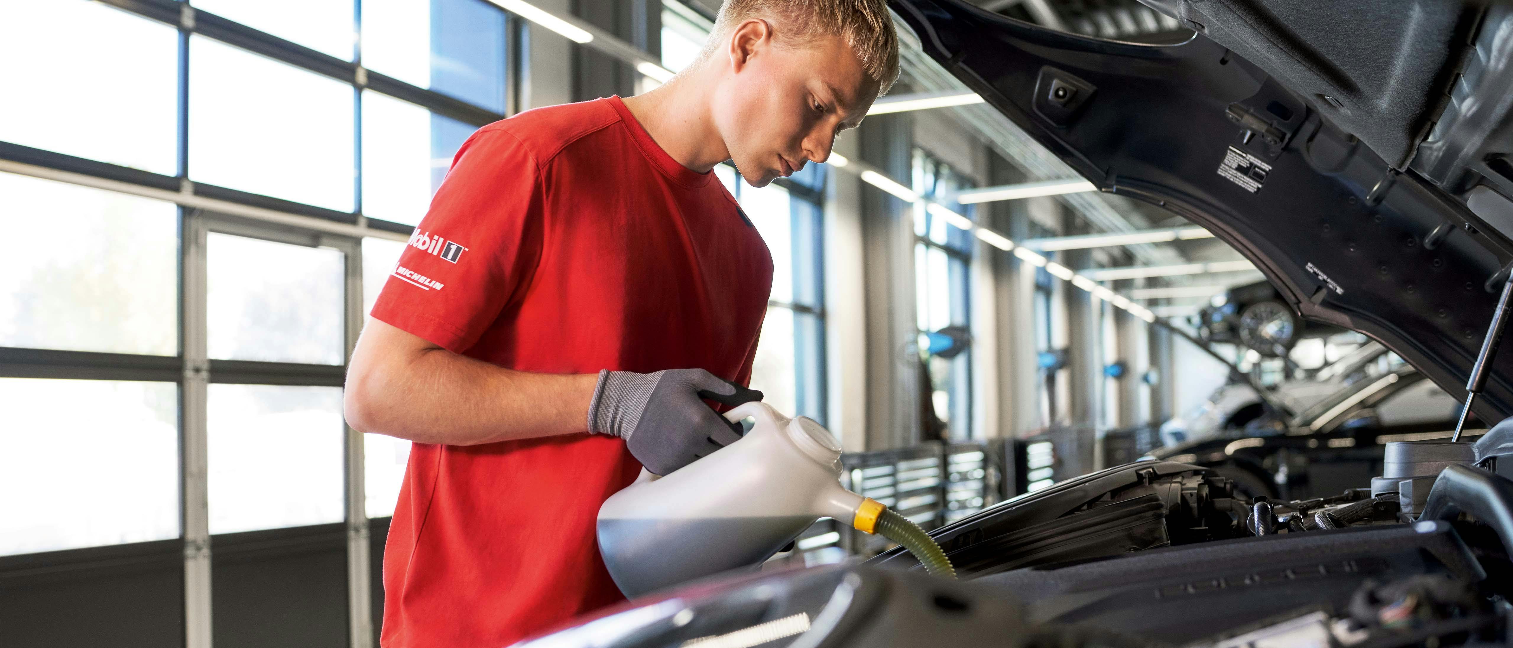 Een man in een rode werkplaats Porsche polo giet vloeistof uit een kan in de motor van een auto. Hij draagt een grijze handschoen en kijkt geconcentreerd naar wat hij doet. De motorkap van de auto is open.