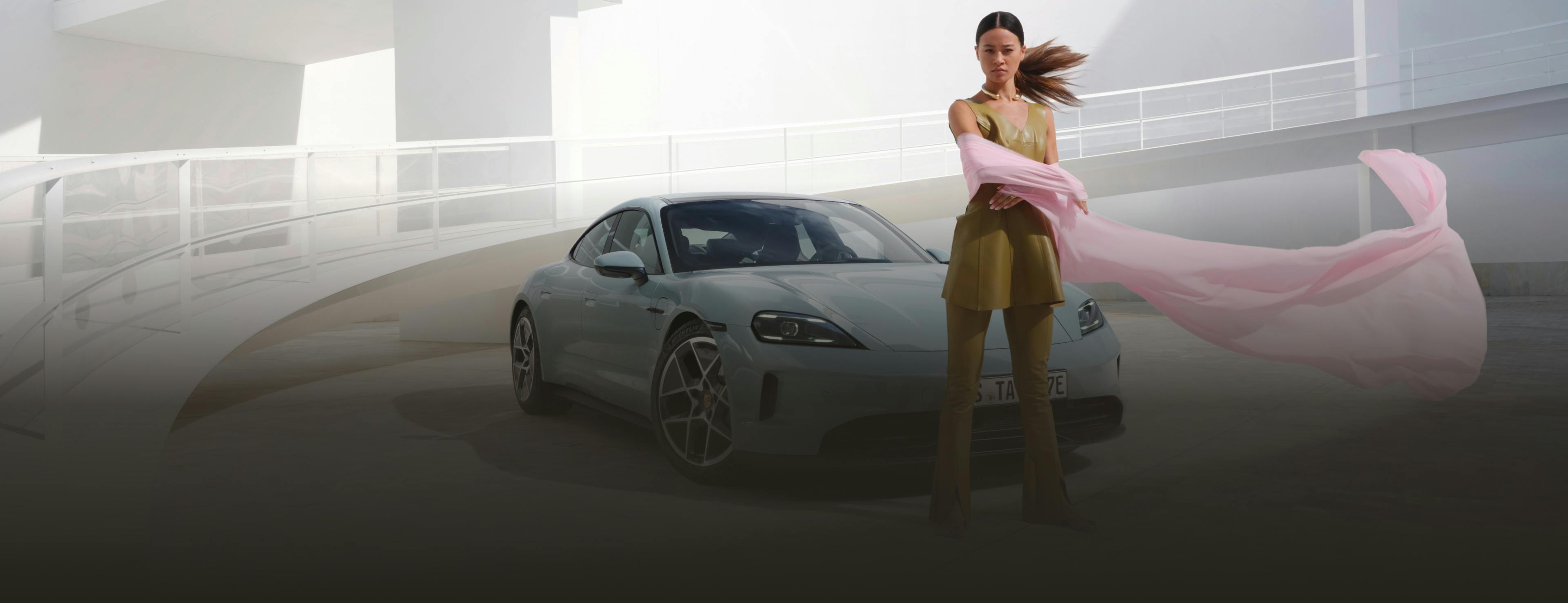 View of the front and side of the Porsche Taycan in Ice Gray Metallic. A woman is standing in the foreground.