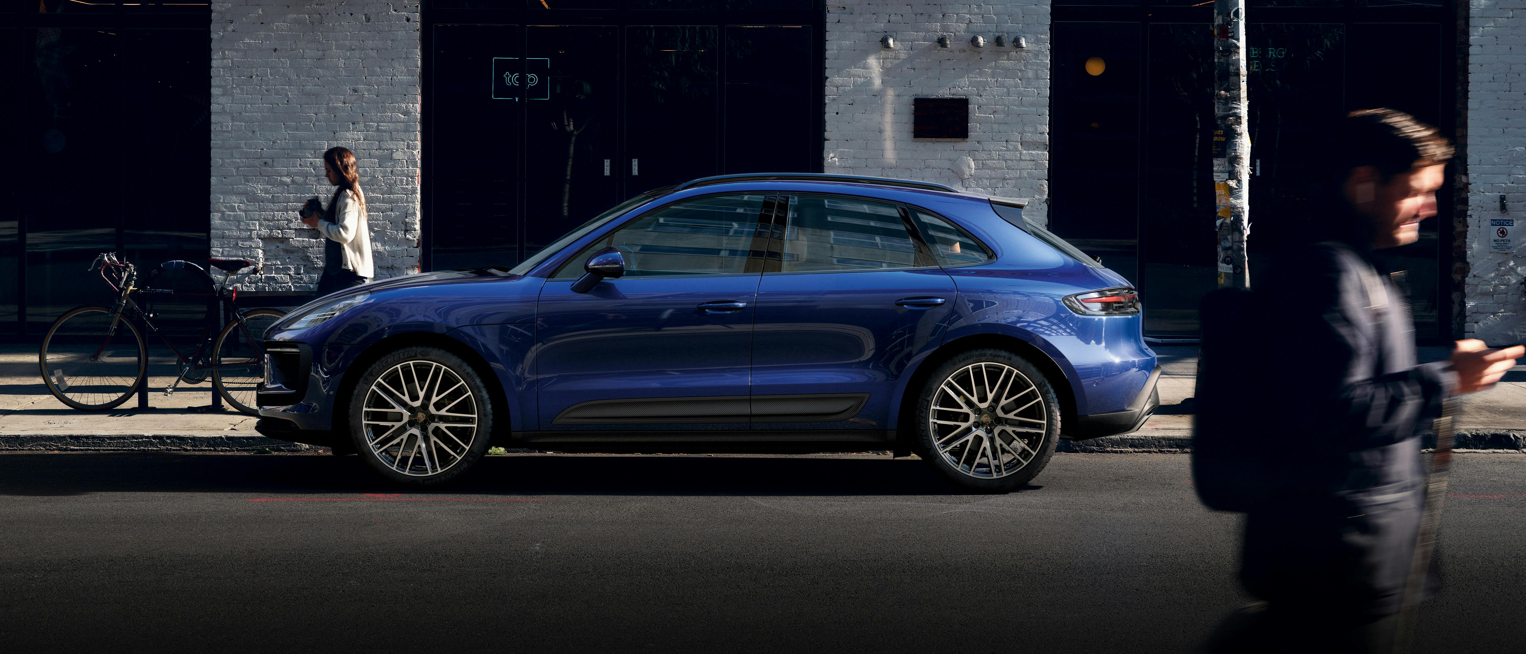 The Macan parked on the road, in front of a white building that looks like a shop, there is a man walking past in the foreground while a woman walking in the opposite direction in the foreground, nearer to the car.