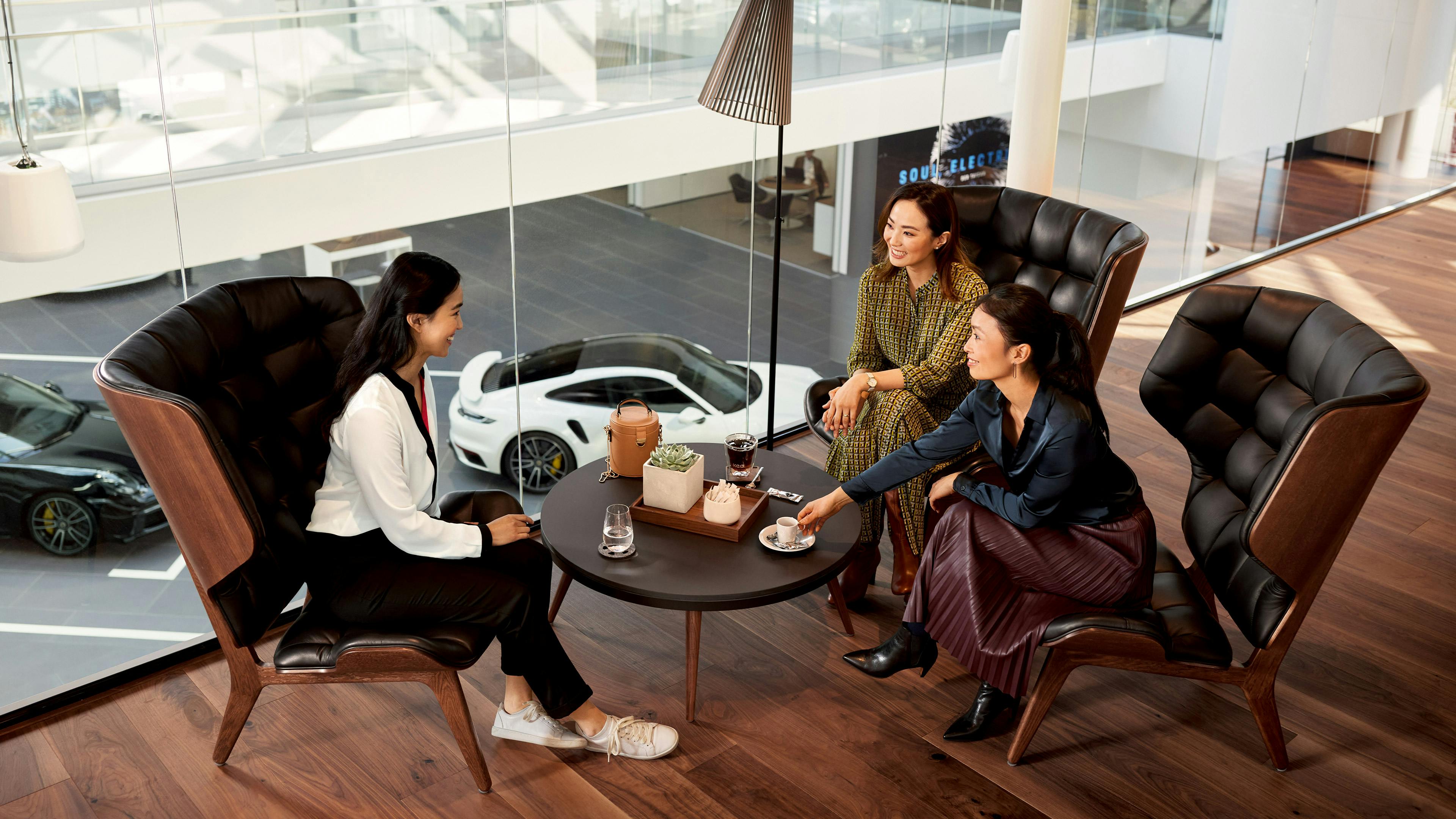Two customers and a Porsche consultant are sitting in a Porsche showroom. Porsche cars can be seen through the window in the background.