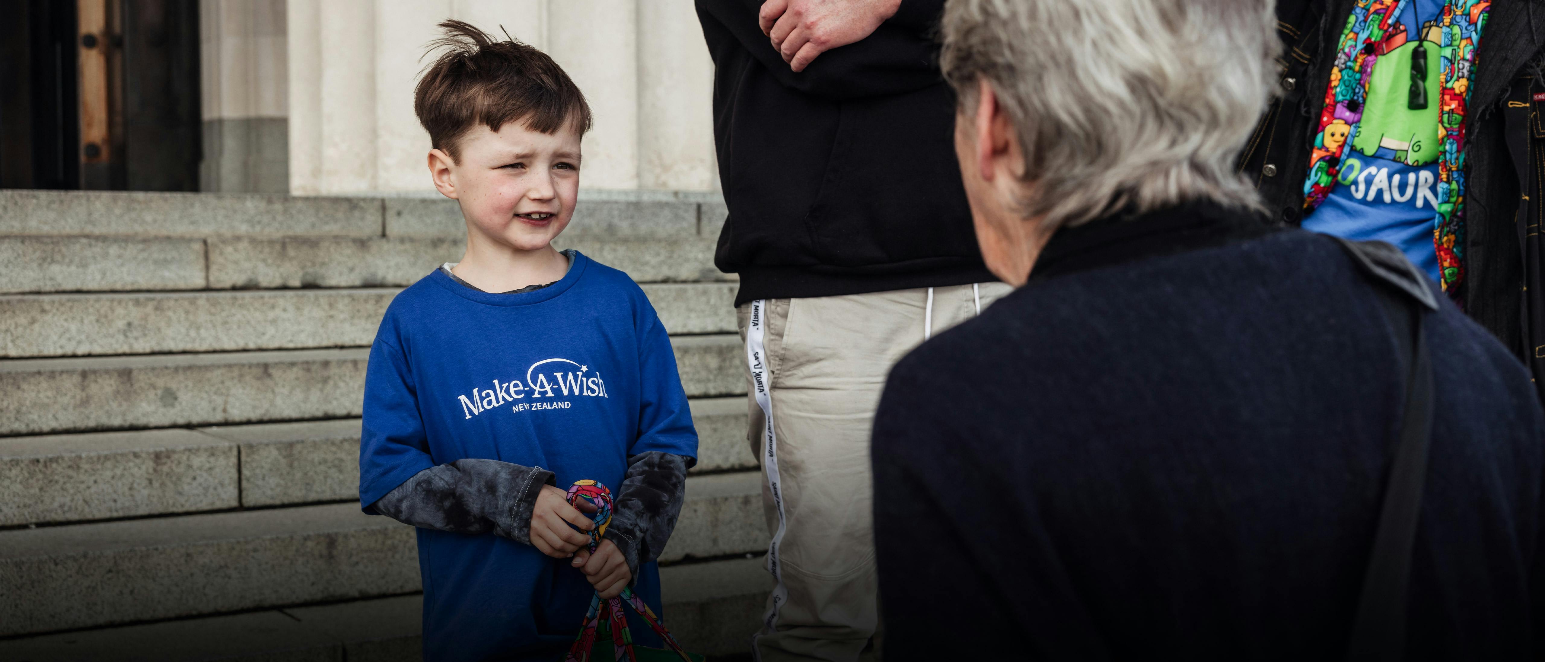 A young child wearing a Make-A-Wish blue long-sleeved shirt stands on stone steps, looking up towards an older adult whose back is to the camera.