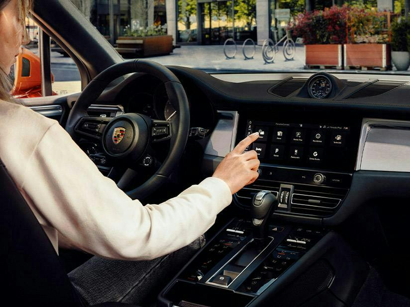 An interior shot from the driver's side of a Porsche, showing a person's hand reaching out to touch the large infotainment screen. The dashboard features a classic analogue clock in the centre and a leather-wrapped steering wheel with the Porsche crest. Through the windshield, a sunlit urban plaza with bike racks and planters is visible