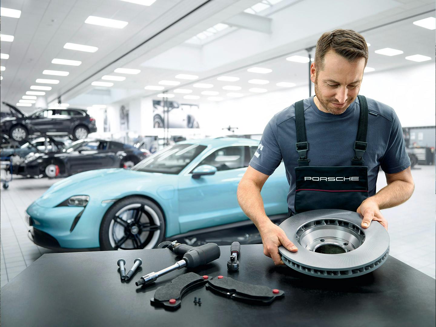 Ein Porsche-Techniker inspiziert eine Bremsscheibe in einer modernen Werkstatt, mit einem türkisfarbenen Porsche Taycan in Hintergrund