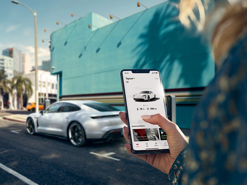 A hand holding a smartphone with the My Porsche App and a white Porsche Taycan in the background.