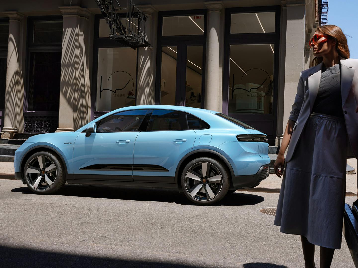 The Macan Electric parked on the side of a road, with a woman leaning on a van in the foreground.