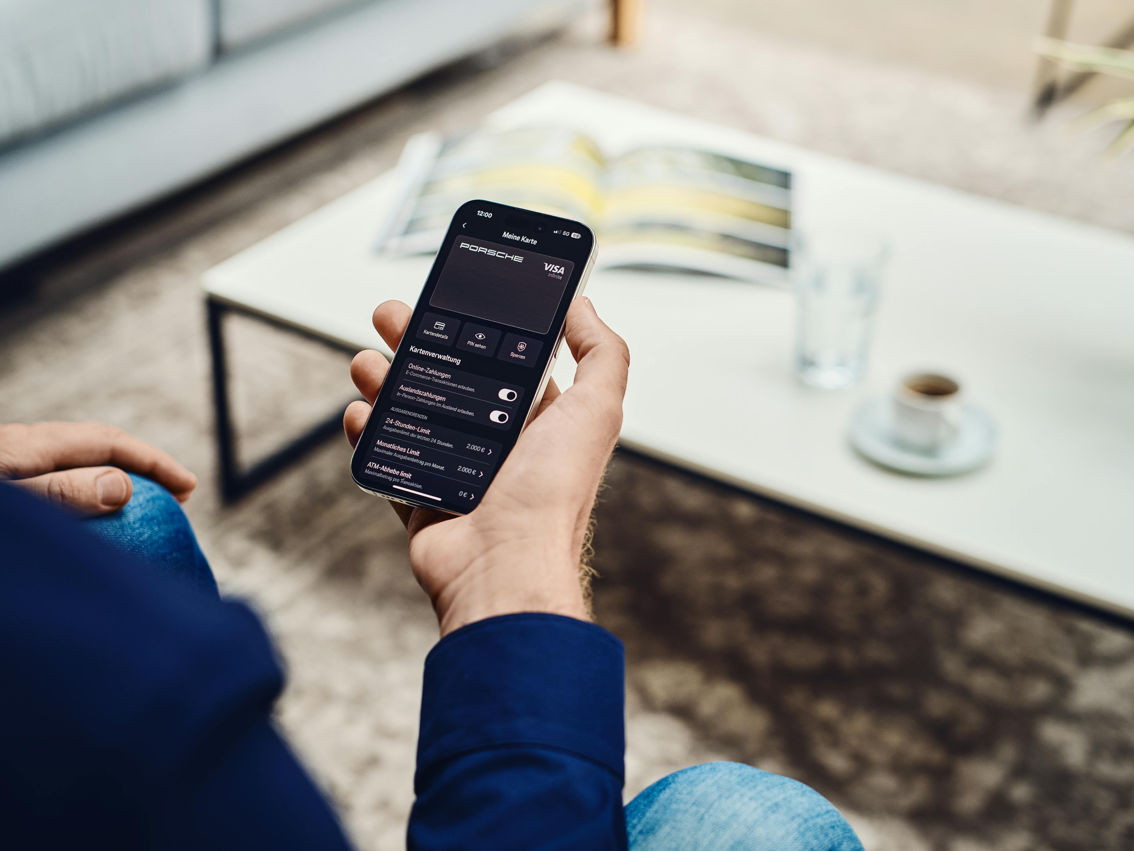 Ein Mann sitzt in einem modernen Wohnzimmer und hält ein Smartphone in der Hand. Auf dem Display ist die Porsche Card Visa innerhalb der Porsche App zu sehen. Im Hintergrund stehen eine Tasse Kaffee, ein Glas Wasser und ein aufgeschlagenes Magazin auf einem Couchtisch.