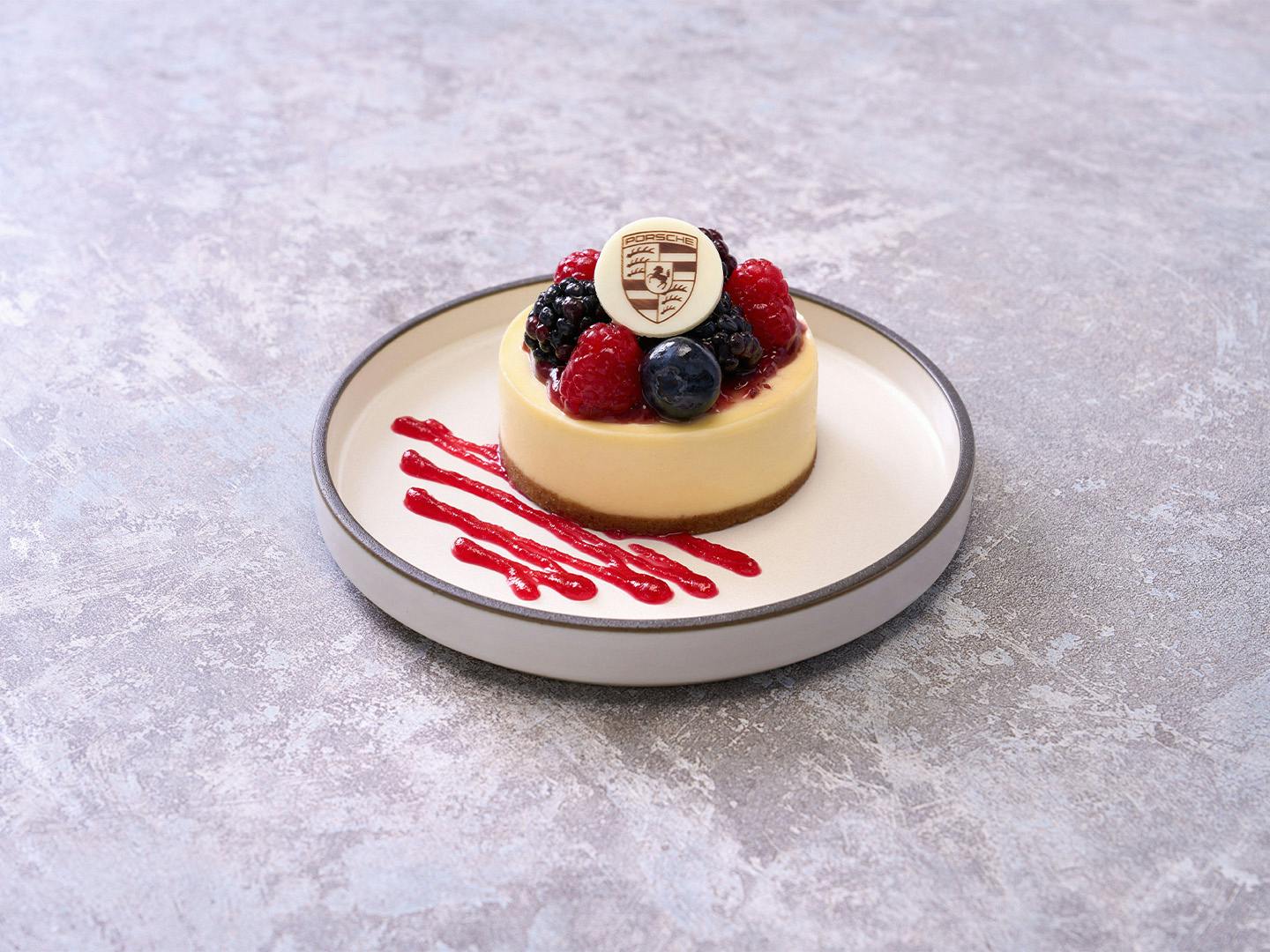 Mini cheesecake with fresh berries and white chocolate Porsche logo on plate with raspberry sauce.