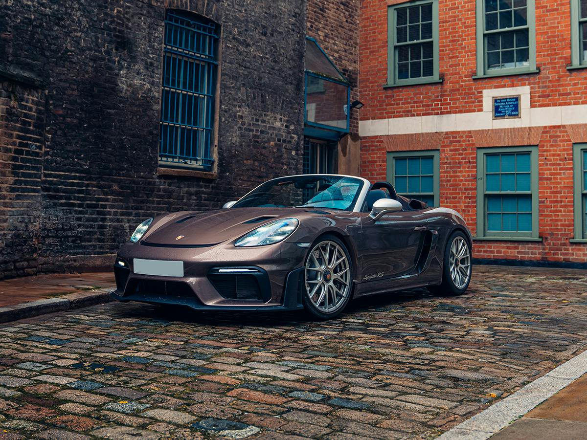 A Porsche 718 Boxster Spyder RS in Paint to Sample Chestnutbrownmetallic parked on a cobblestone street between historic brick buildings.