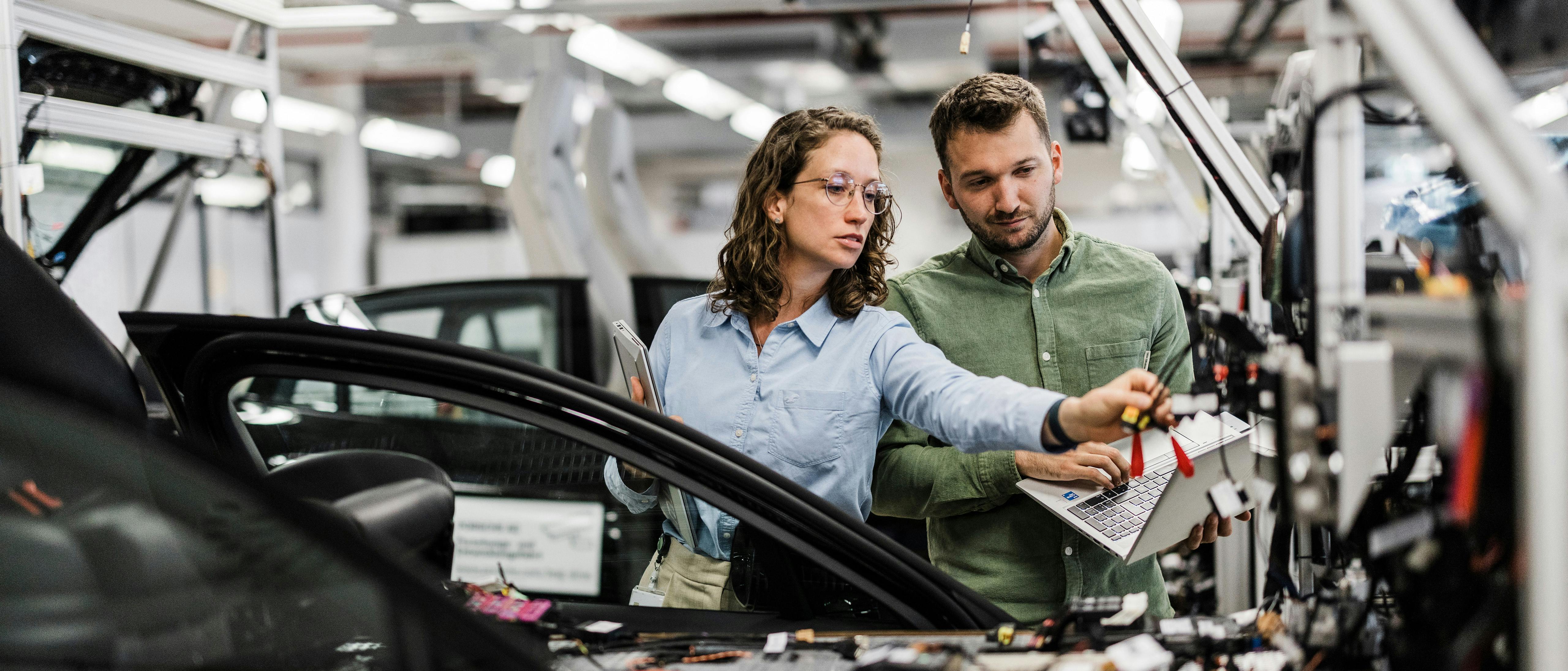 Zwei Personen arbeiten gemeinsam an einem Fahrzeug in einer modernen Produktionshalle, stehen neben einem geöffneten Auto mit sichtbaren Kabeln und Bauteilen und nutzen Werkzeuge sowie Unterlagen.