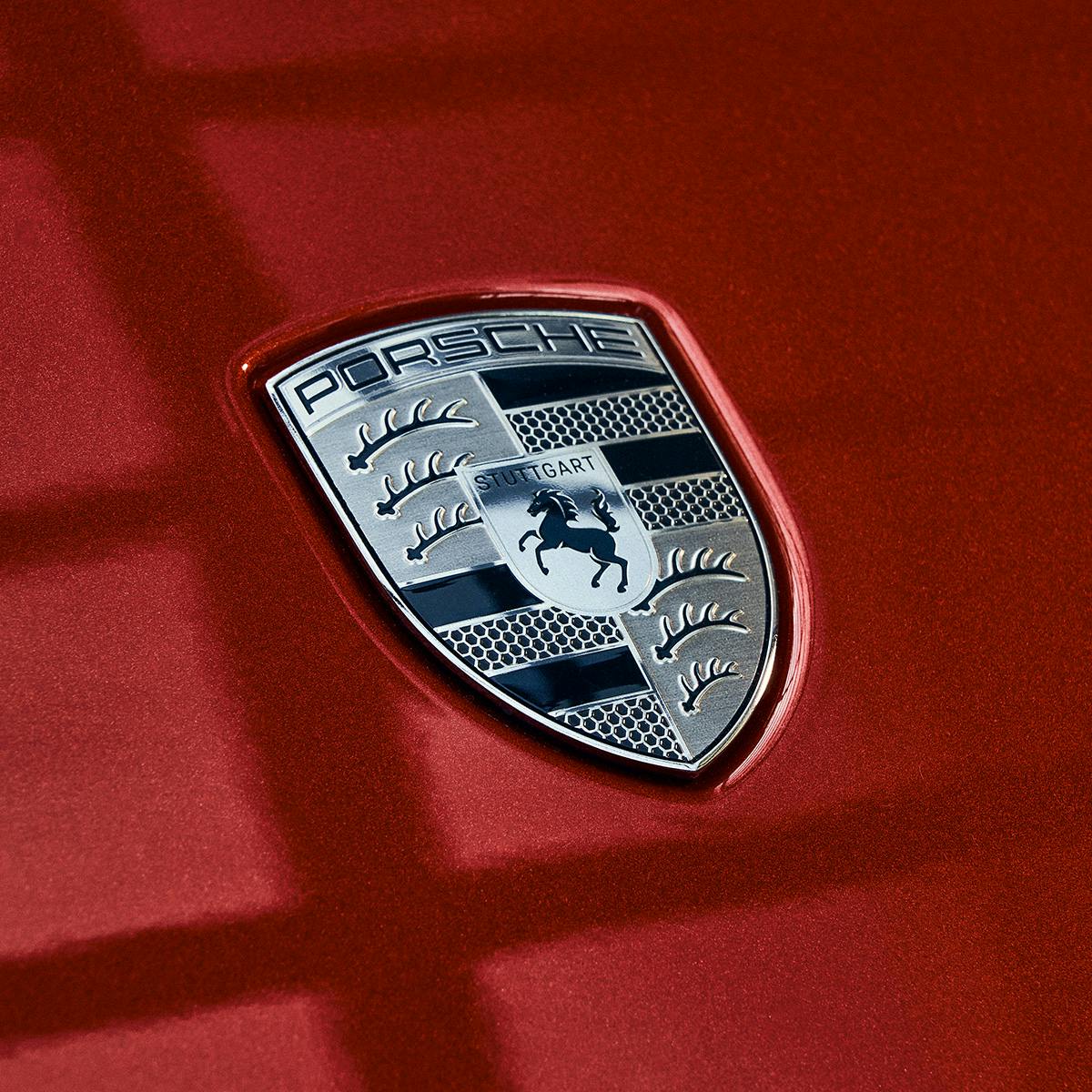 A close-up of the Porsche logo on a red car. The emblem features the word "Porsche" at the top, a black prancing horse in the center.