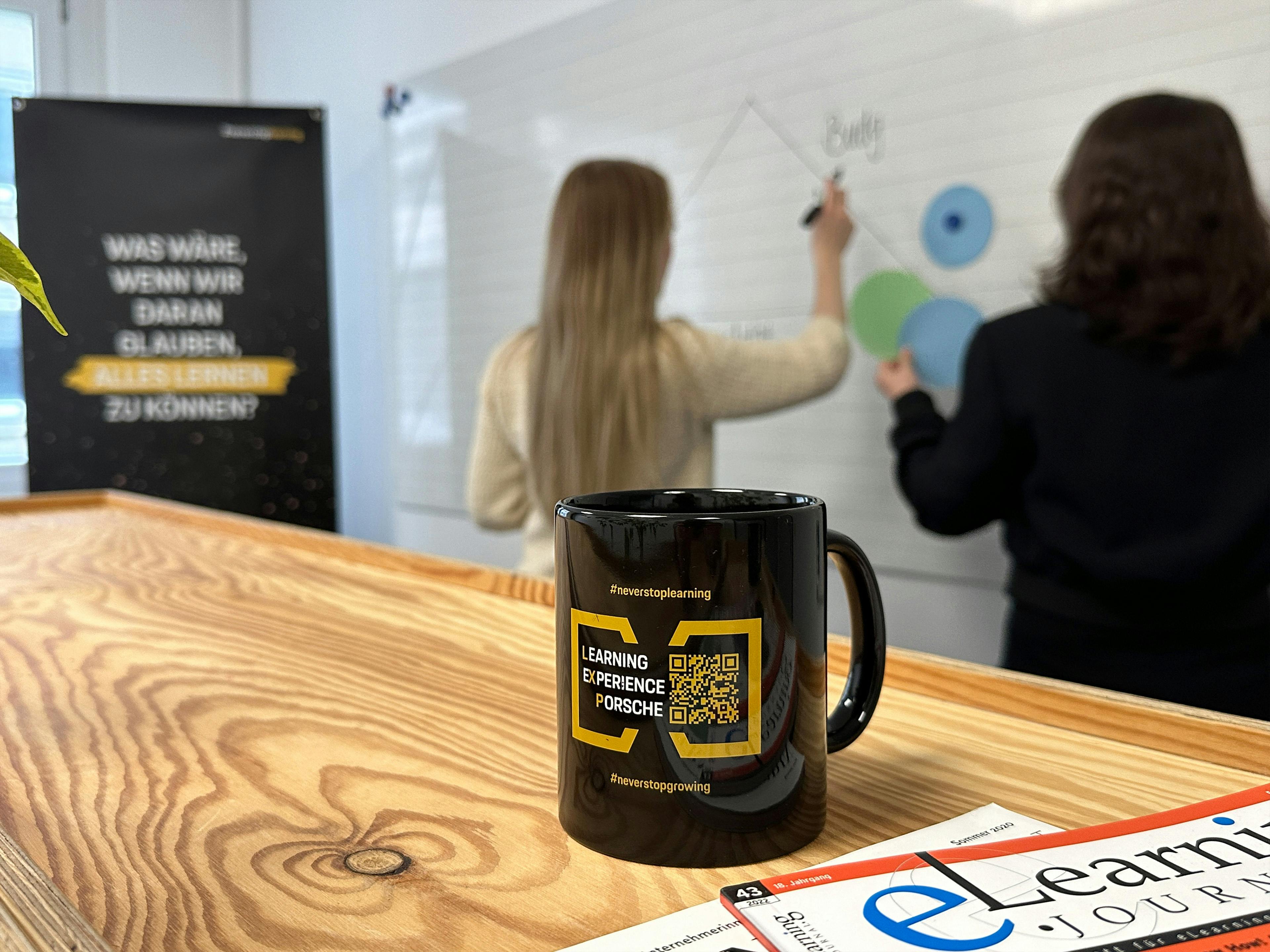 Zwei Mitarbeiterinnen stehen an einem Whiteboard, im Vordergrund ist eine Tasse zu sehen, die Mitarbeiter motiviert neues zu entdecken.