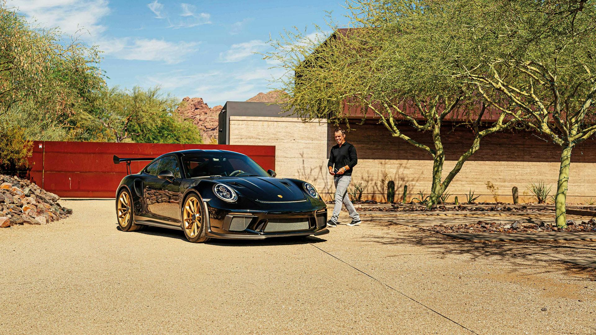 Paul Casey läuft lächelnd auf einen schwarzen Porsche mit goldenen Felgen zu