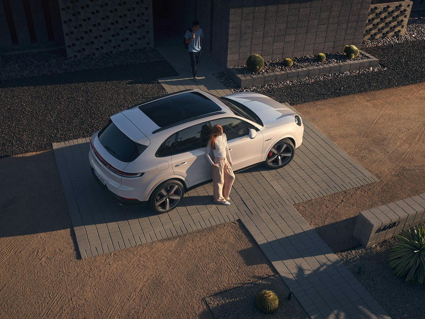Luftaufnahme eines modernen Wohnhauses. Ein weißer Porsche Cayenne steht in der gepflasterten Einfahrt, eine Person neben der geöffneten Fahrertür.